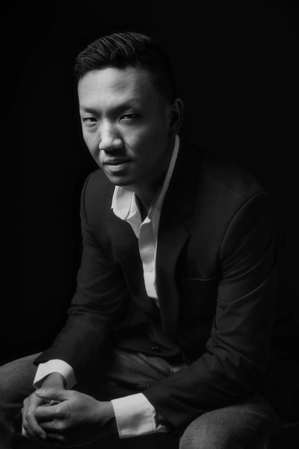 Black and white portrait of a young Asian man in a suit and white shirt, looking into the camera with a serious expression against a dark background.