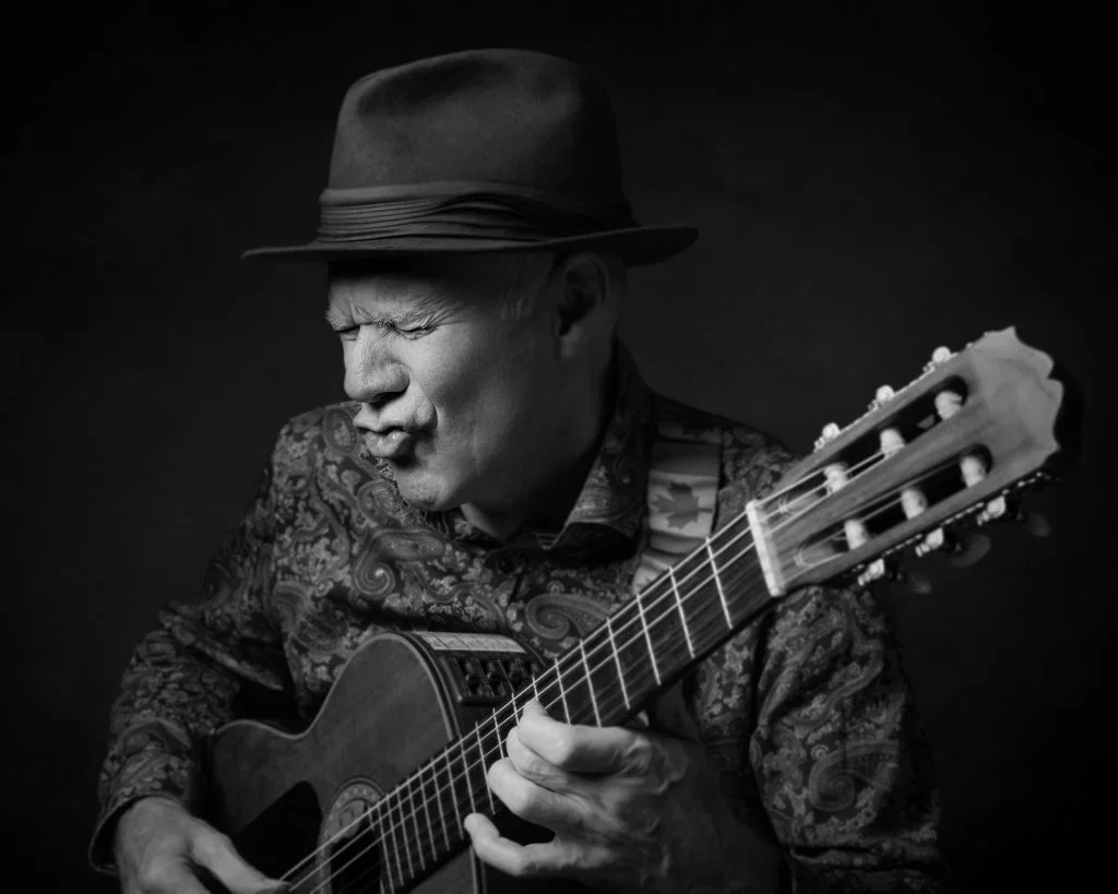 Man wearing a hat playing an acoustic guitar, with a passionate expression, in black and white.