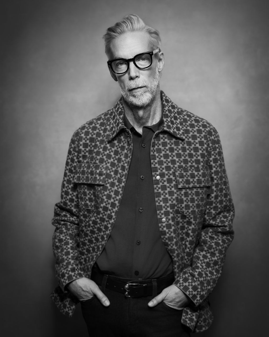 Black and white portrait of a stylish man with a grey beard, black eyeglasses, and a patterned jacket.