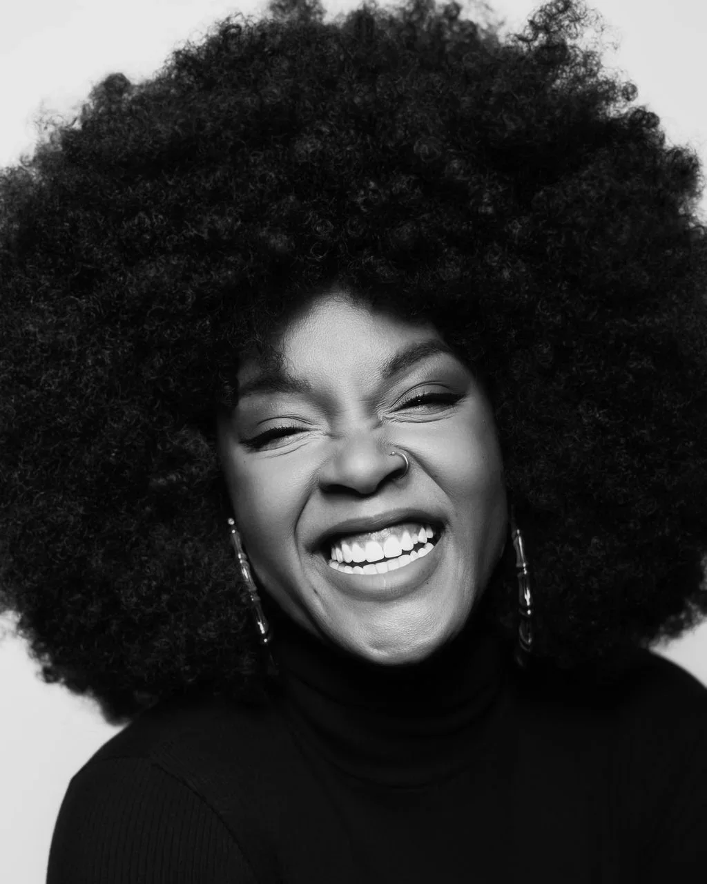 Black and white portrait of a black woman with voluminous curly afro, smiling broadly with her eyes closed, wearing a black turtleneck.
