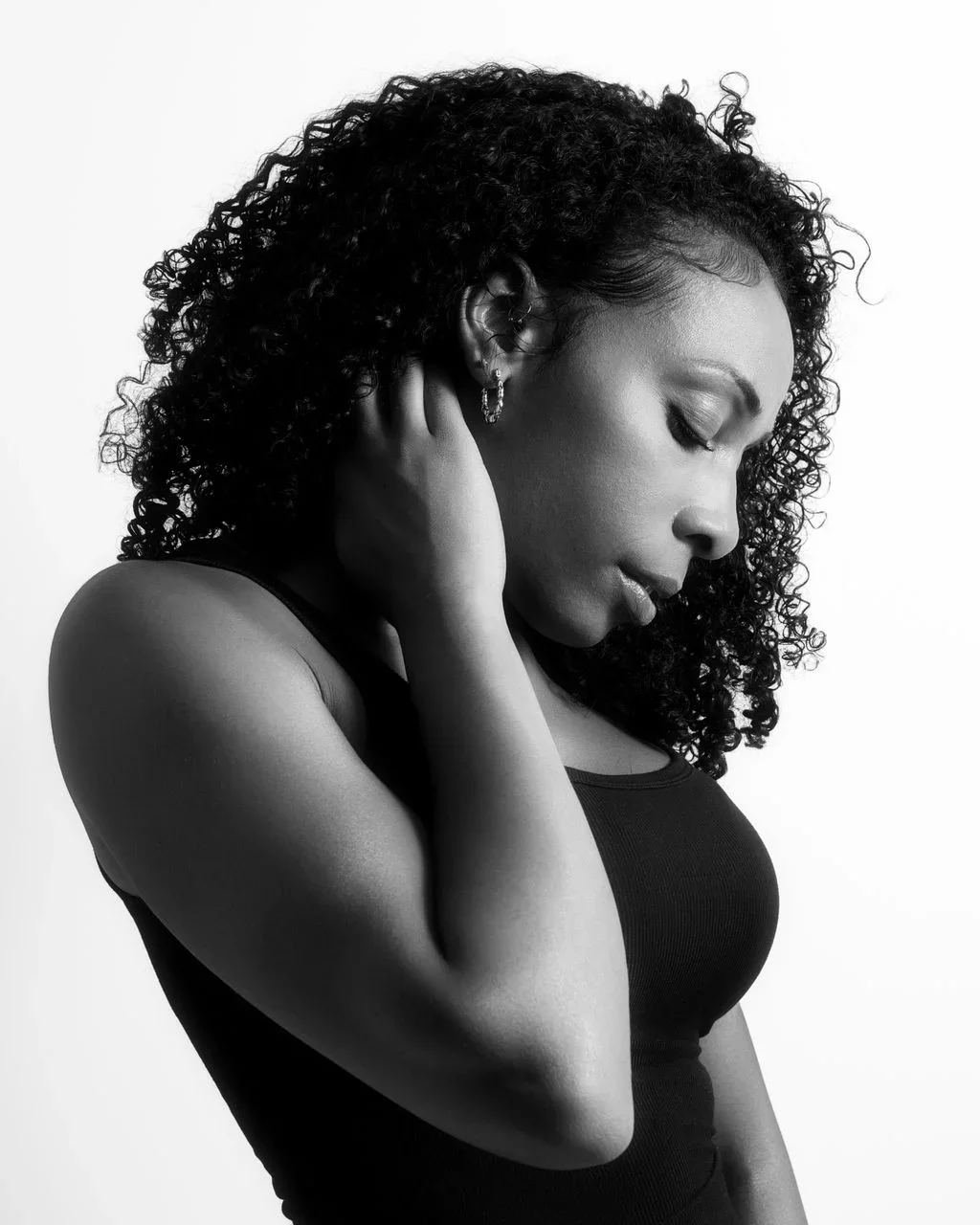 Black and white profile photo of a young black woman with curly hair, eyes closed, touching her neck with one hand, wearing hoop earrings and a sleeveless top.
