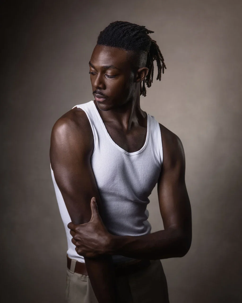 Portrait of a young black man with dreadlocks, wearing a sleeveless shirt, looking down and holding his arm.