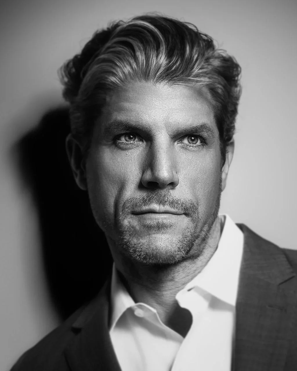 Black and white portrait of a man with grey hair and beard stubble, wearing a crisp white shirt under a blazer leaning against a wall.