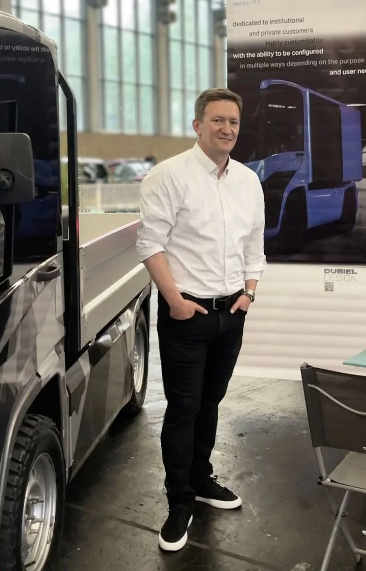 A man in a white shirt and black pants standing indoors near a display of a blue autonomous delivery truck, with large windows and a poster in the background.