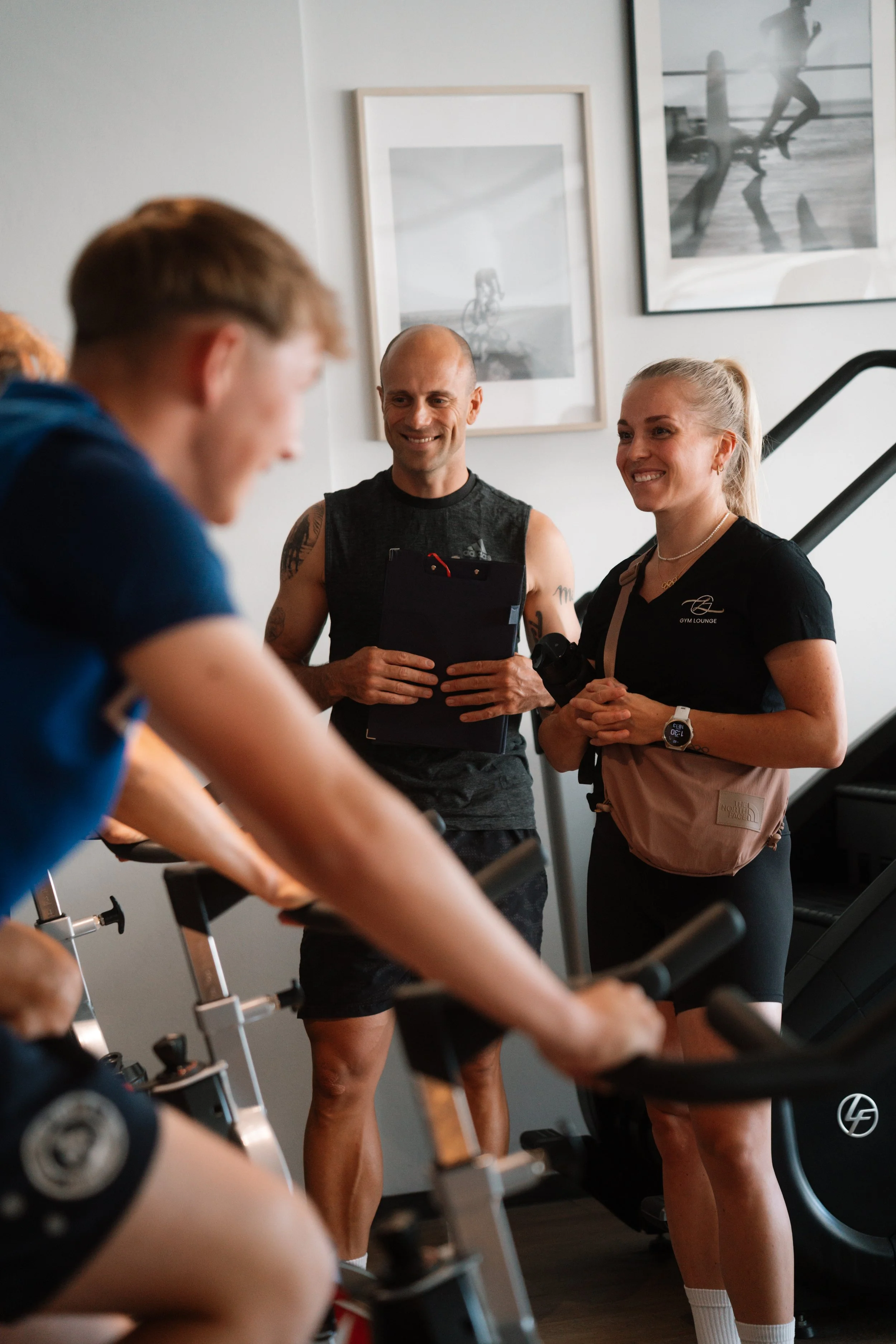 Zwei Menschen, ein Mann und eine Frau, stehen in einem Fitnessstudio, lachen und unterhalten sich, während eine andere Person auf einem Heimtrainer trainiert.