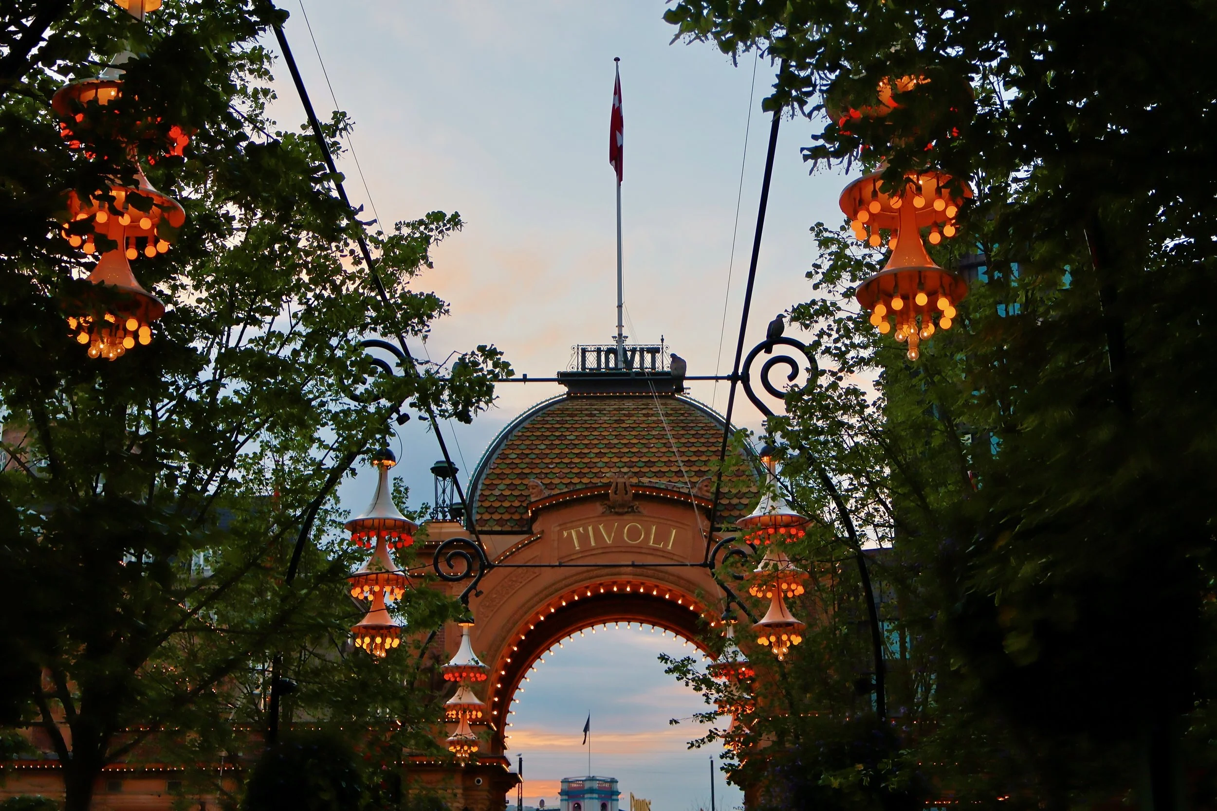 The entrance arch to Tivoli amusement park with ornate hanging lights, trees on either side, and a Swiss flag on a pole at the top of the arch during twilight.