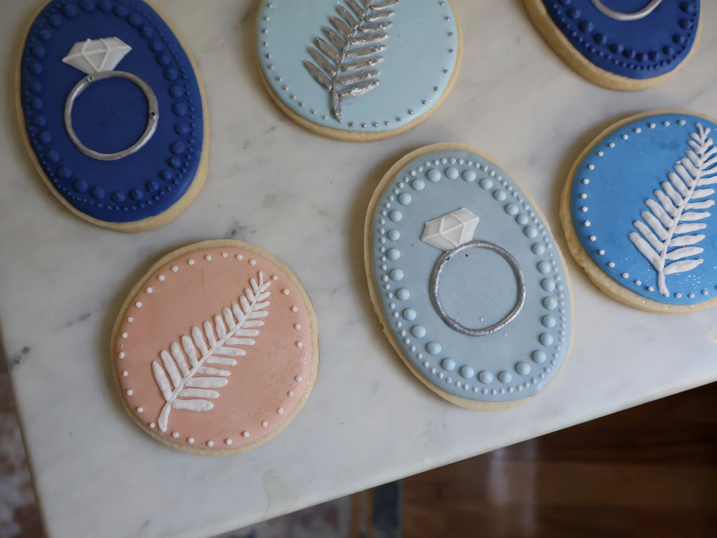 Decorated cookies with pastel blue, pink, and dark blue icing featuring fern leaves, pearls, and a silver ring with a diamond.