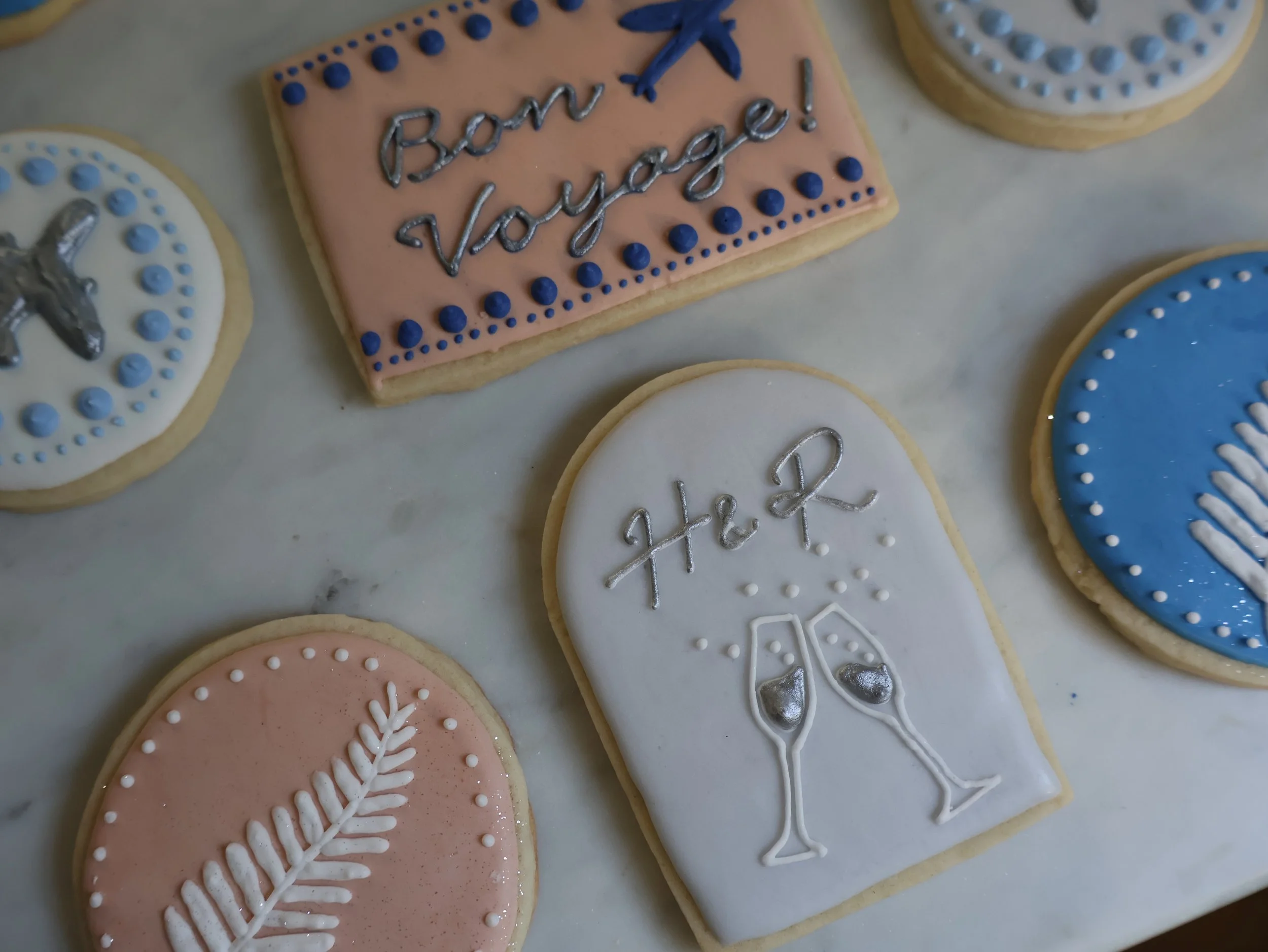 Decorated cookies, including one with the words 'Bouwt Voyage!', and others with various patterns and designs, some with silver and pastel icing, on a white surface.