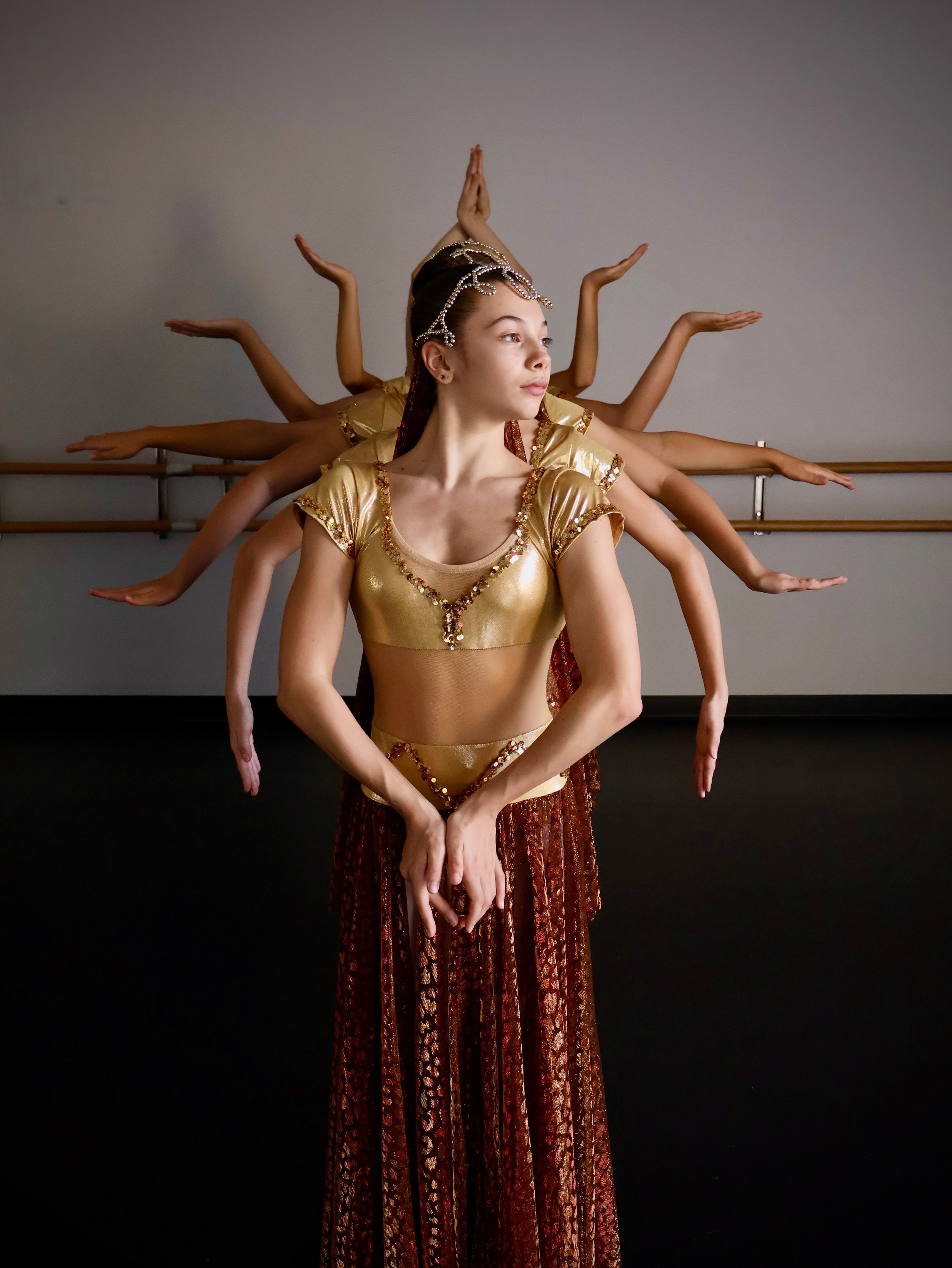 Young female ballet dancer in gold costume with multiple arms, posing in a dance studio.