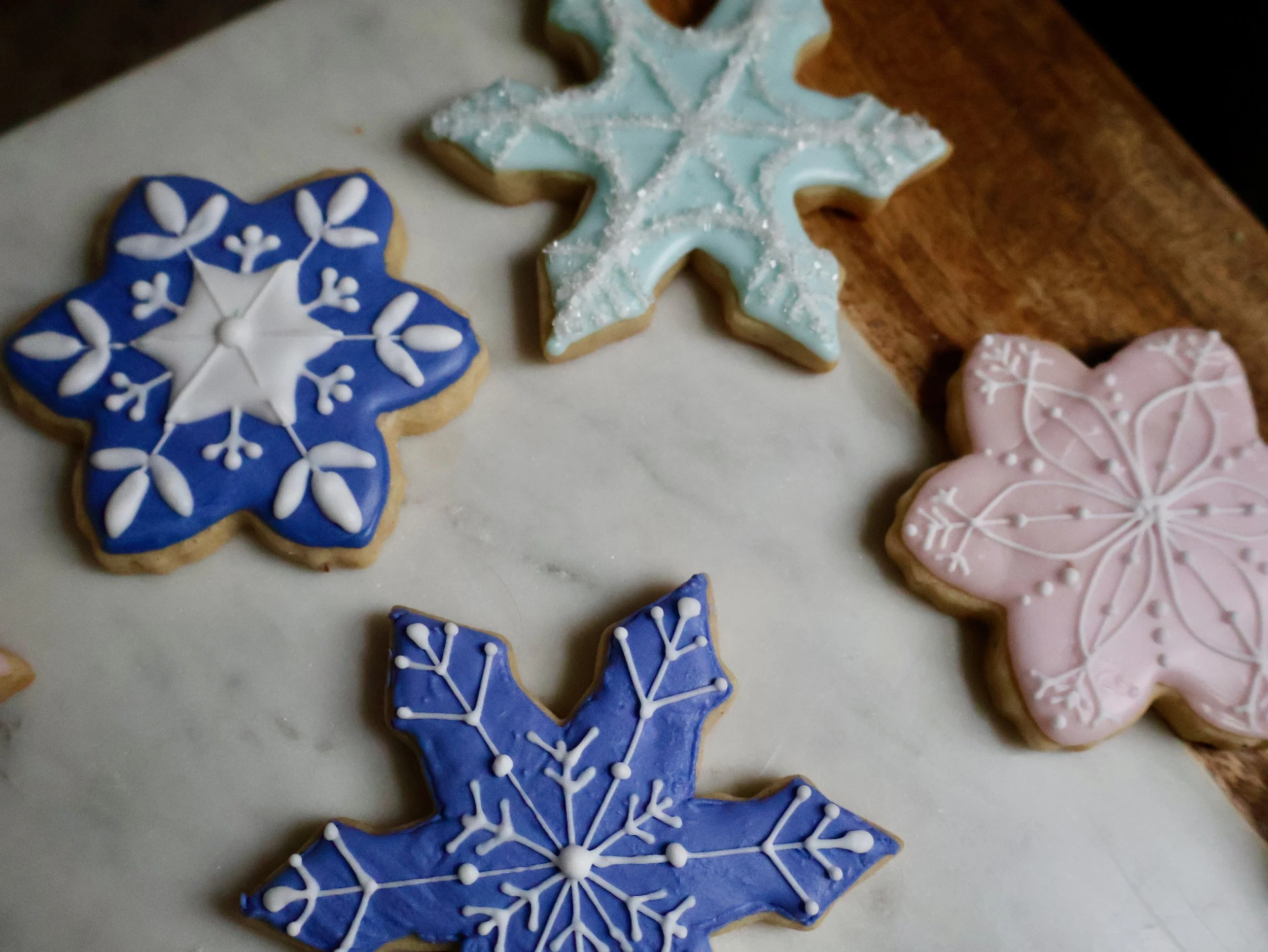 Decorated Christmas cookies in snowflake shapes with blue, pink, and light blue icing, some with white snowflake patterns, on a marble surface near a wooden cutting board.