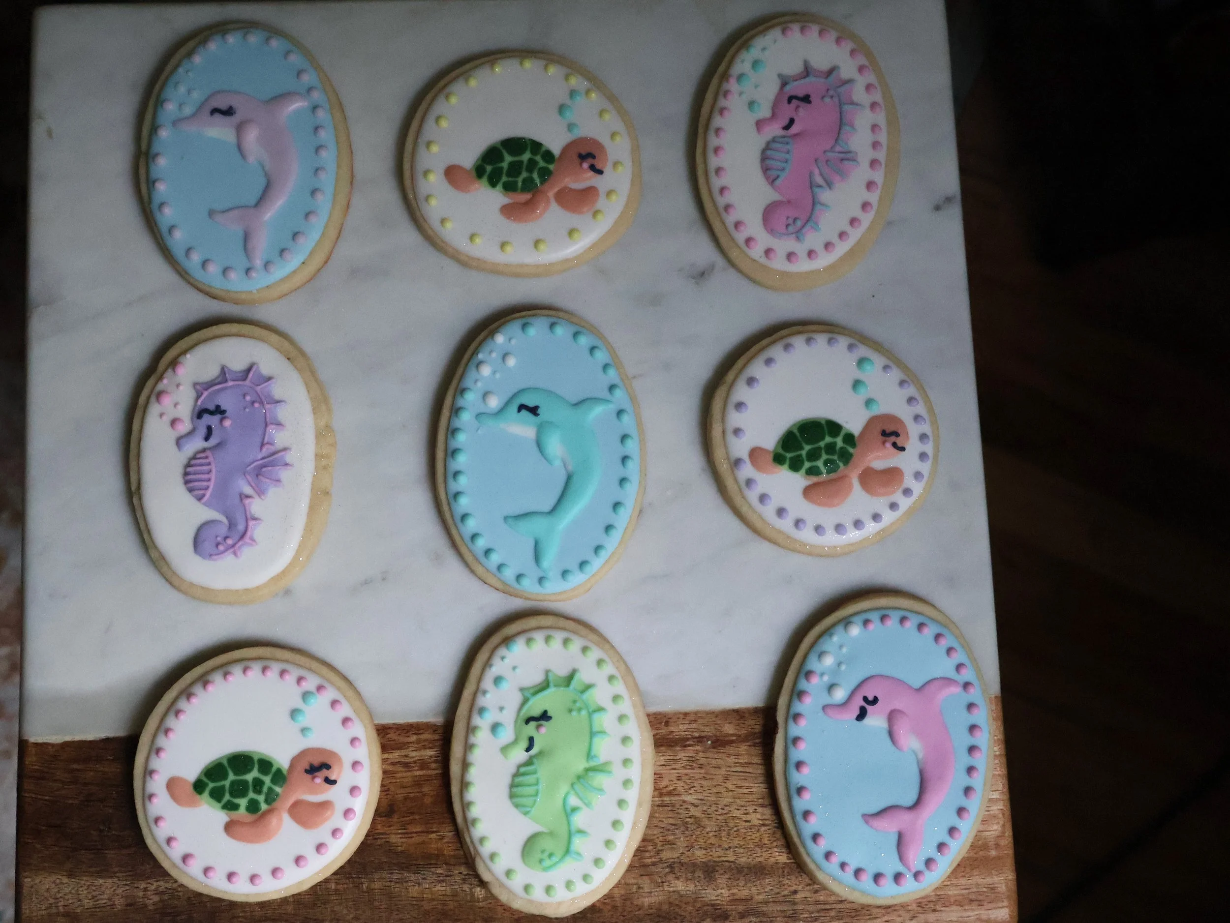 Decorated cookies with sea animal designs, including dolphins, turtles, and seahorses, on a marble and wooden surface.