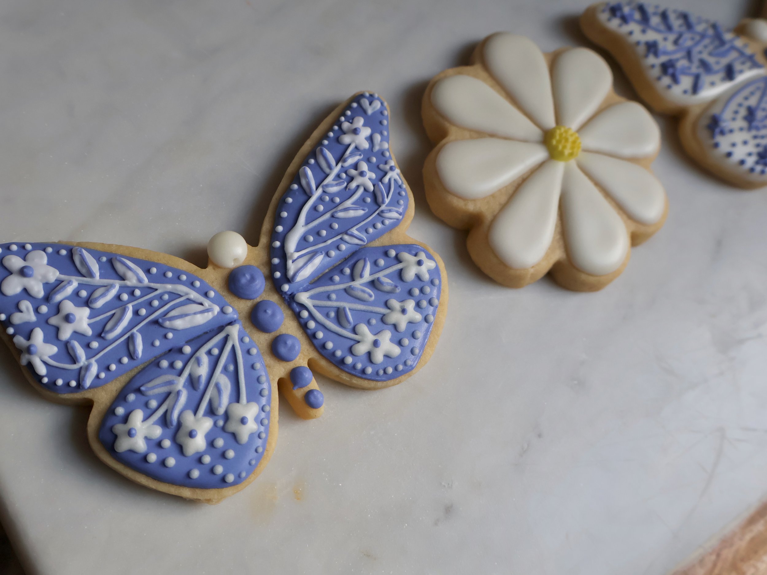 Decorated cookies on white marble surface, including a butterfly-shaped cookie with blue and white icing and floral details, and a white flower-shaped cookie with yellow center.