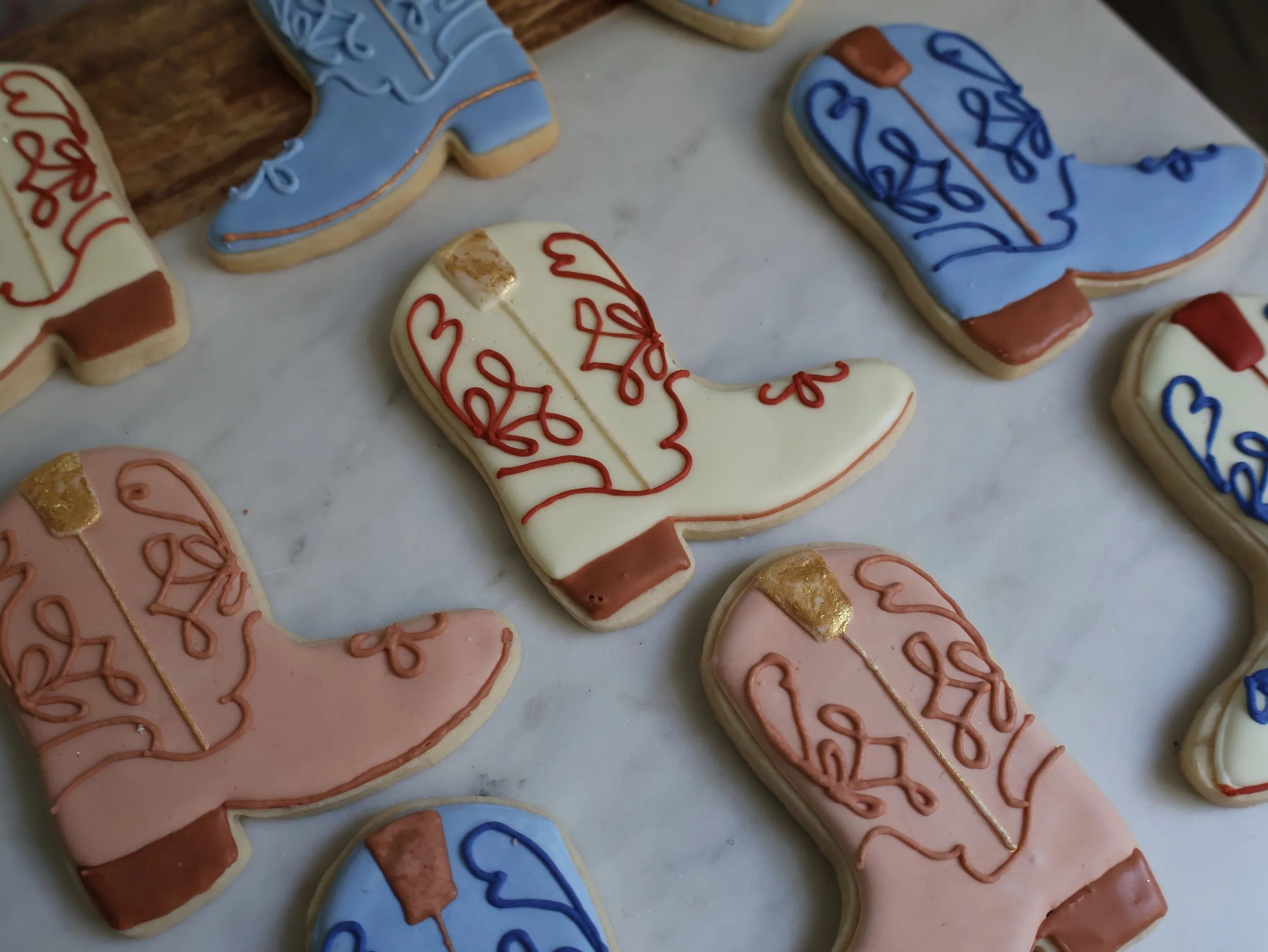 Decorated cookies shaped like snow boots with pink, blue, white, and peach icing with red and gold piping decorations, arranged on a marble surface.