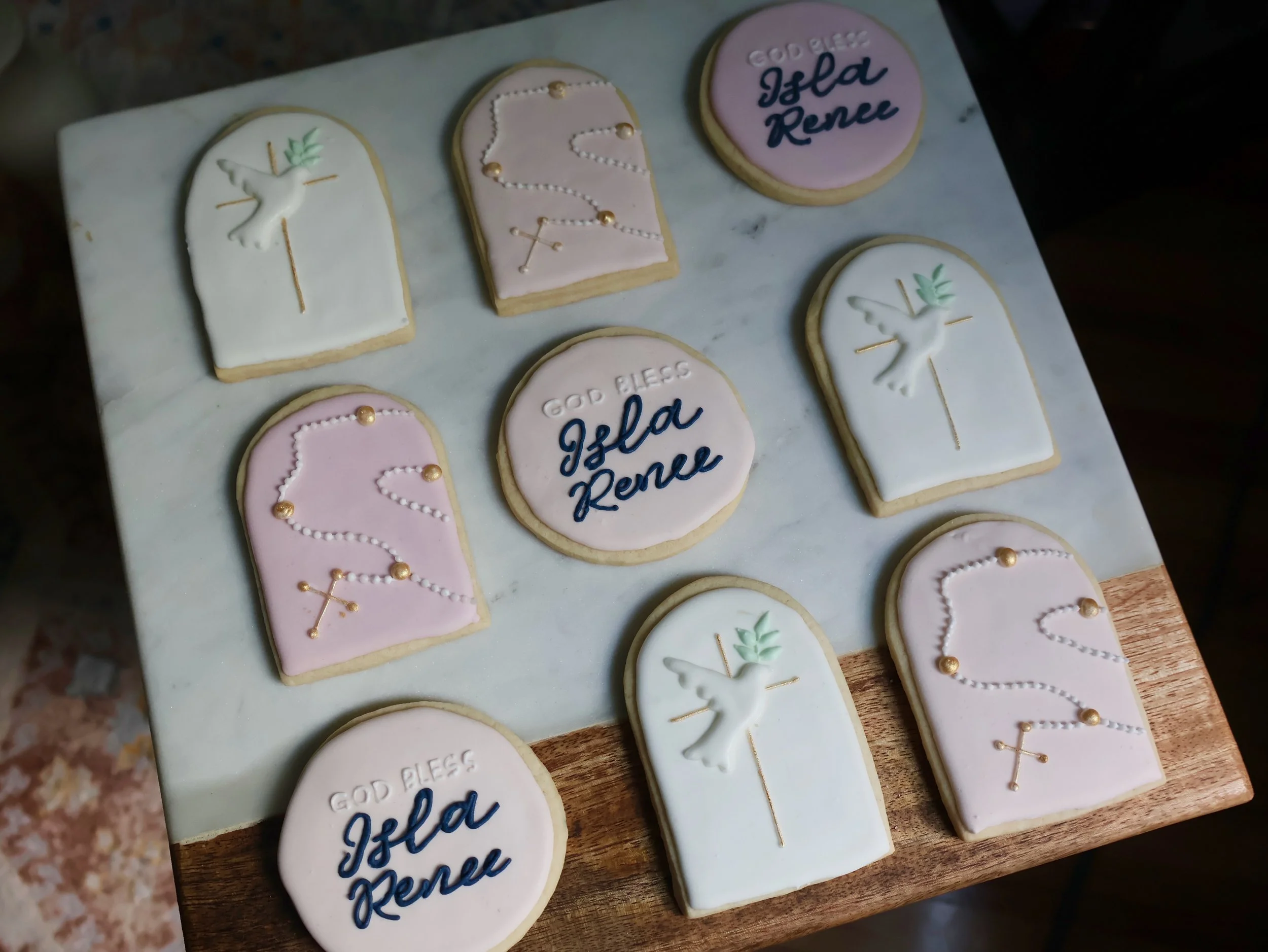 Decorative cookies on a board with Christian symbols and greeting messages. The cookies are in the shape of tombstones and round medallions, decorated with white, pink, and cream icing. Some cookies display a dove and a rosary, while others have insc