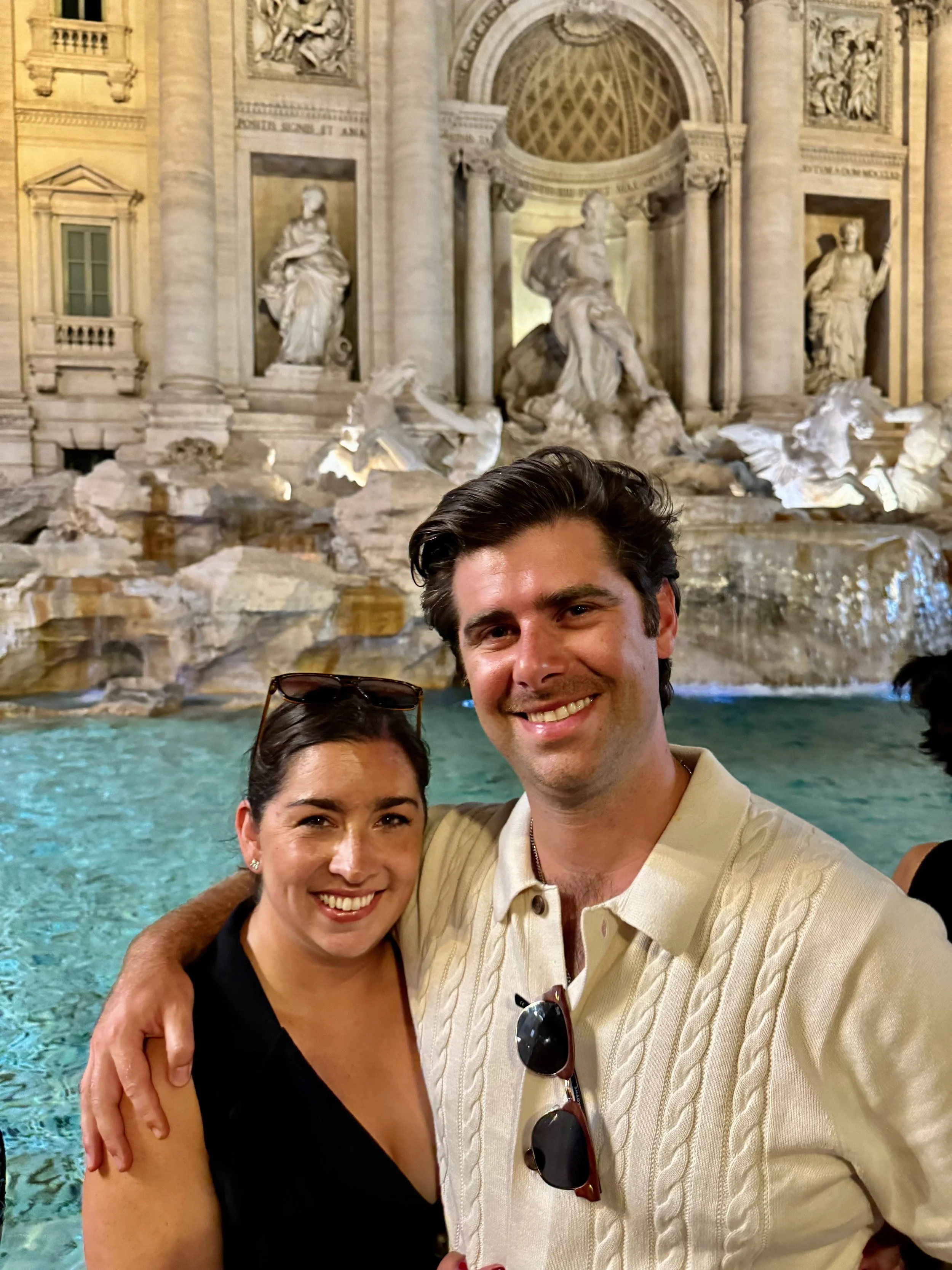 Two smiling people, a woman and a man, embracing each other in front of the Trevi Fountain in Rome, Italy, during the evening.