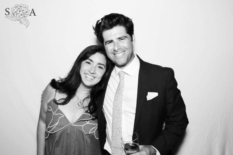 A smiling man and woman posing together at an event, the man holding a glass of wine, both dressed in formal attire, standing close to each other in front of a plain backdrop.
