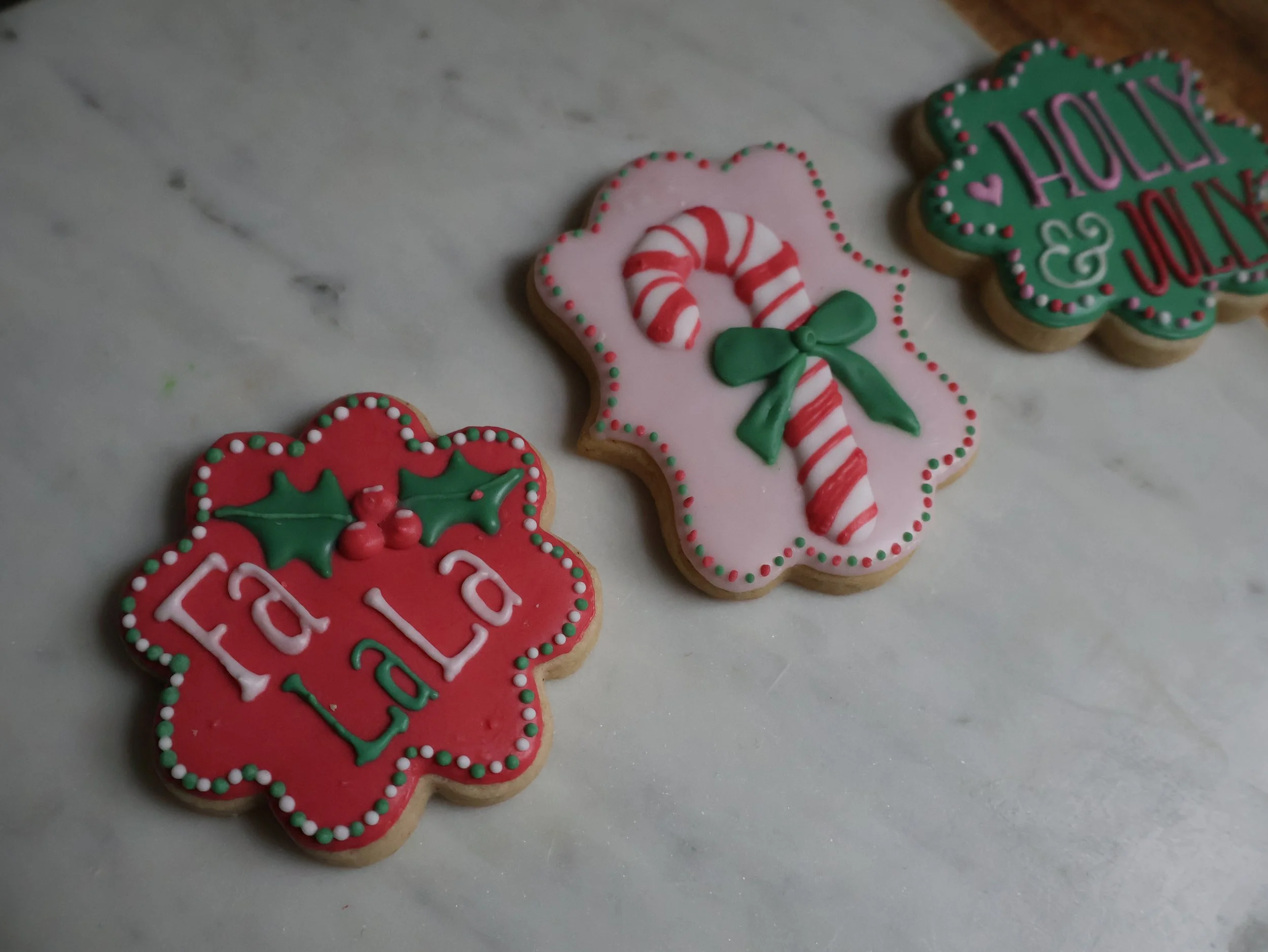 Decorated Christmas cookies on a white surface, including one with HOLLY & JOLLY, another with a candy cane, and a third with a Christmas greeting.