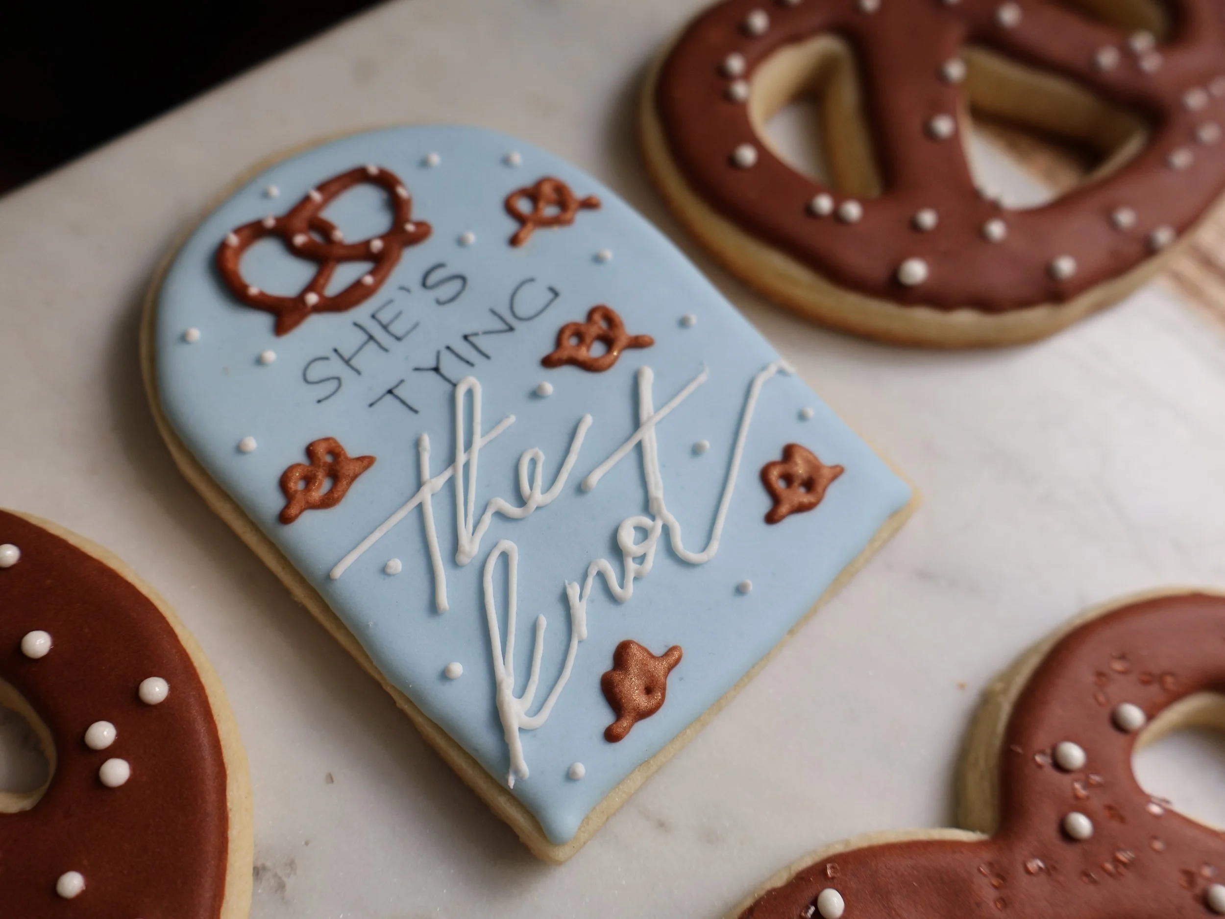 Decorated cookies with Christmas theme, including one blue rectangular cookie with white and brown icing that reads 'She's trying to hide' with pretzel and bow decorations, and round cookies with brown icing and white dot decorations.