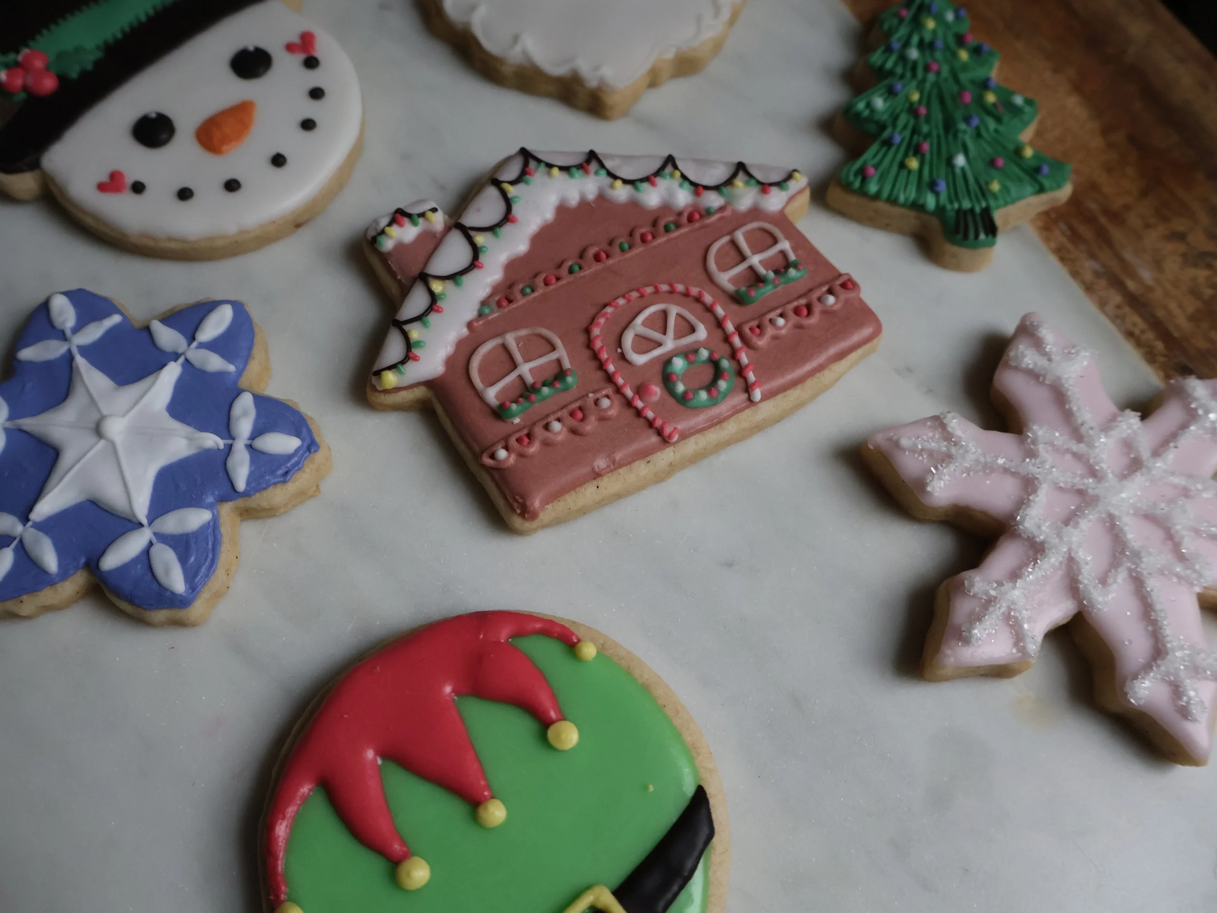 Decorated Christmas cookies shaped like a snowman, house, Christmas tree, snowflake, elf hat, and star, displayed on a white surface.