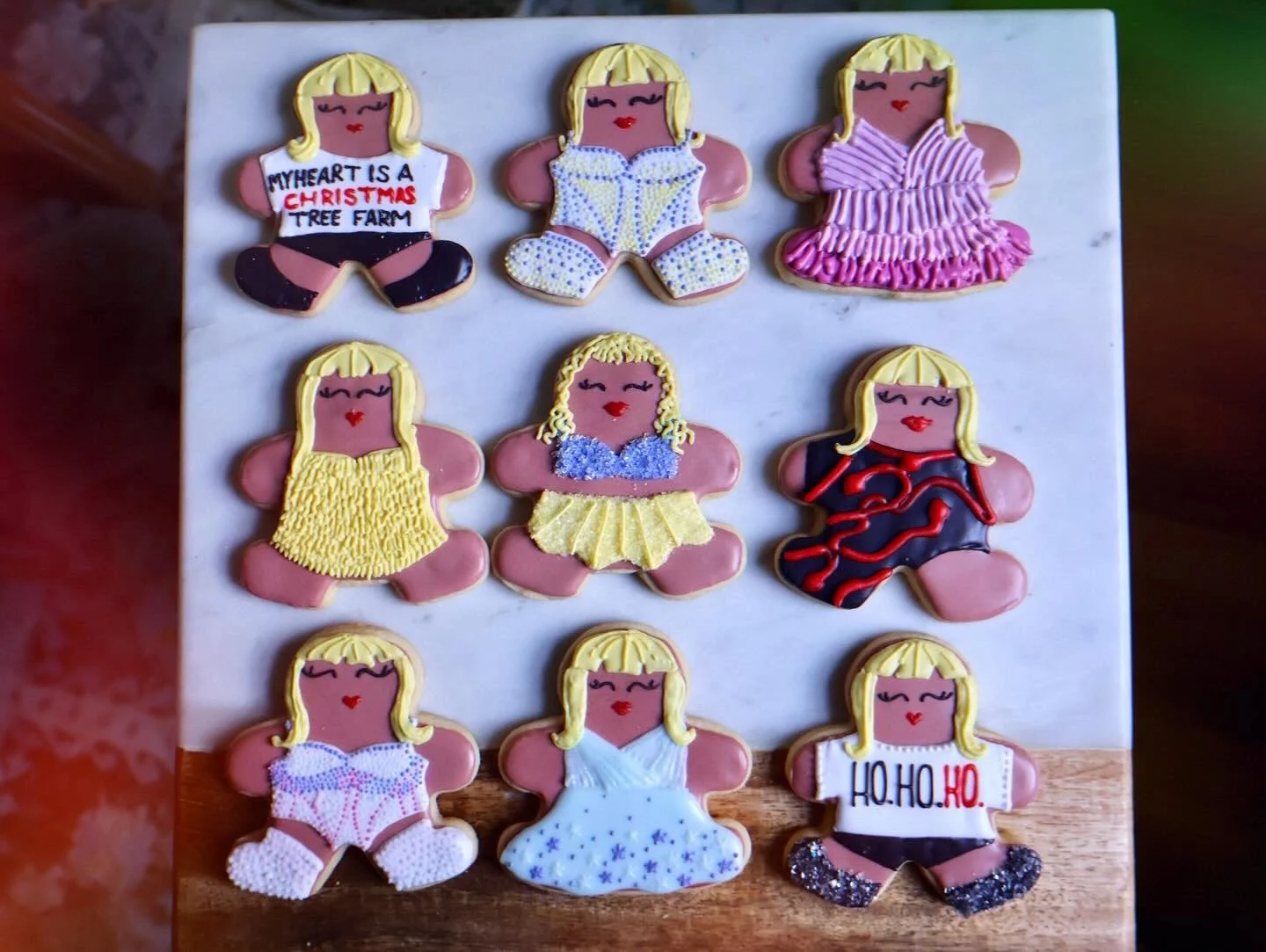 Decorated gingerbread cookies shaped like girls with blonde hair, colorful outfits, and festive details, arranged on a white and wooden surface.