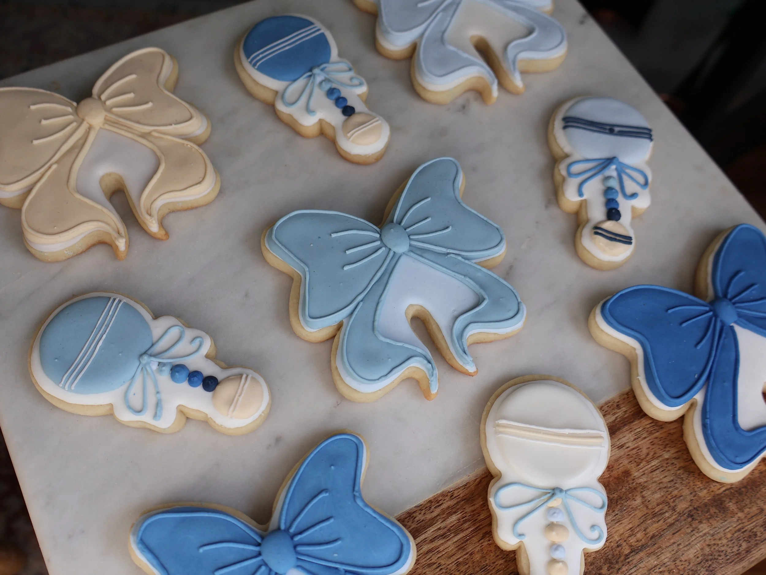 Cookies decorated with blue, white, and beige icing in butterfly and candy shape on a marble and wooden surface.