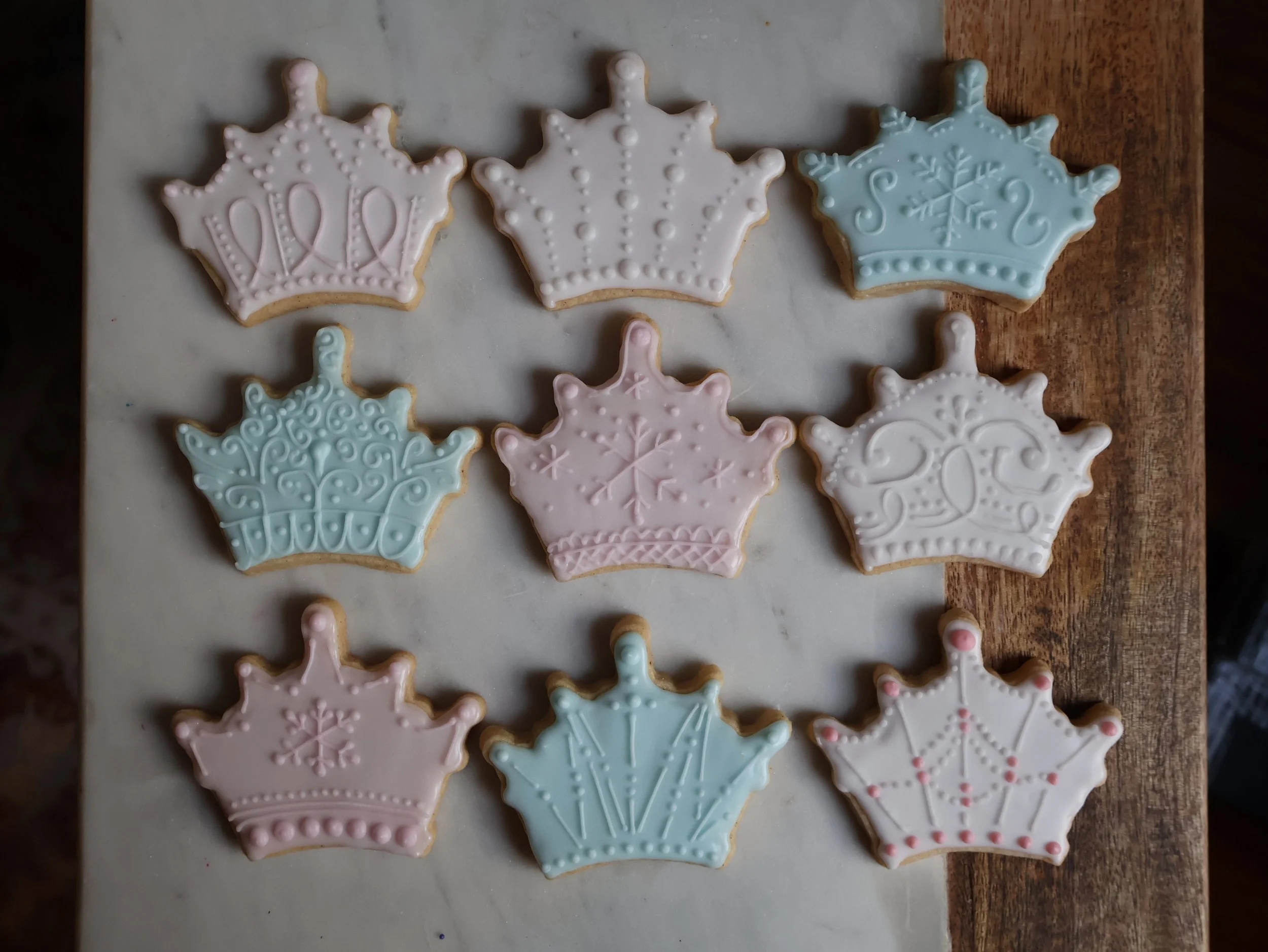 Decorated crown-shaped cookies in pastel pink, blue, and white icing, arranged on a marble surface.