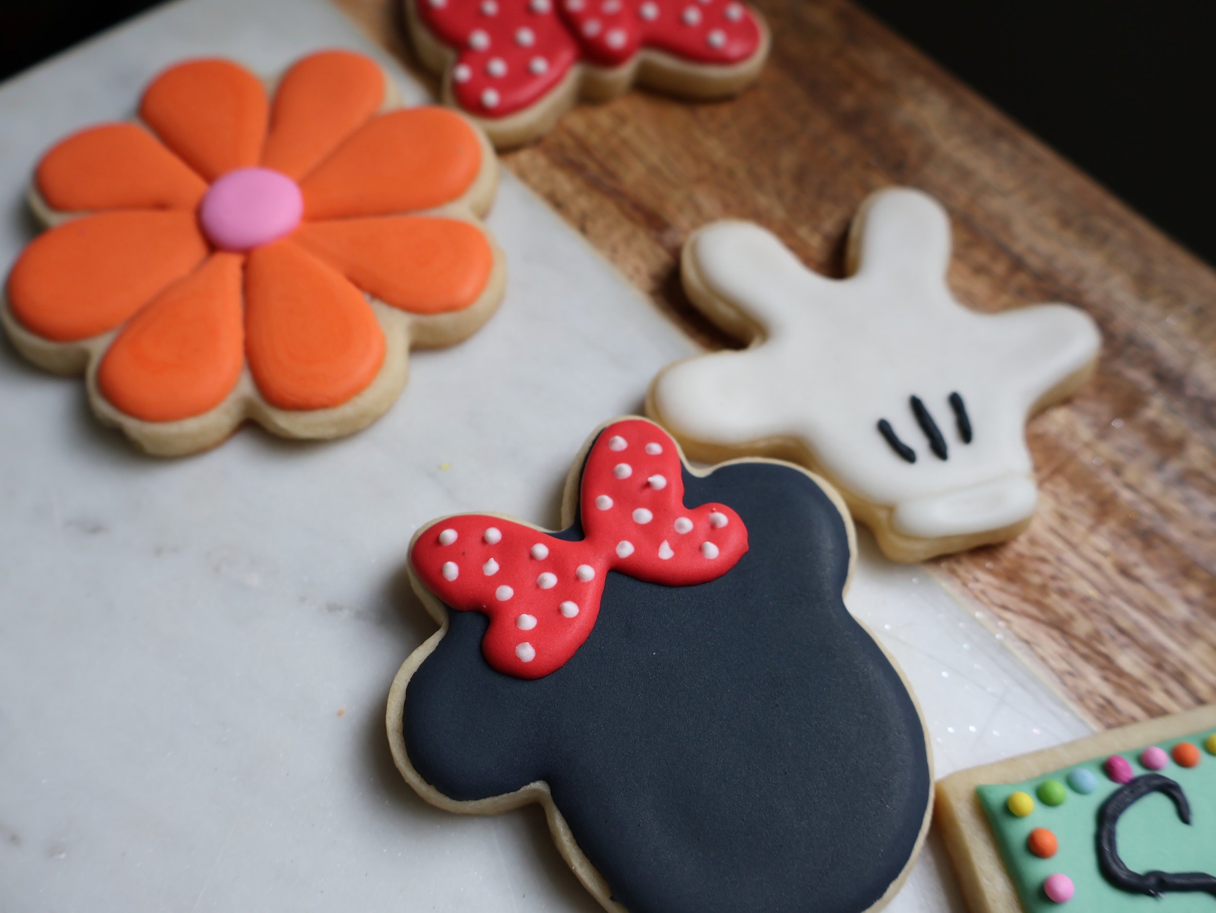 Assorted decorated cookies on a wooden surface, including a black mouse-shaped cookie with a red bow, a white hand-shaped cookie with black lines, a large orange flower-shaped cookie, a red flower-shaped cookie with white polka dots, and a green rect