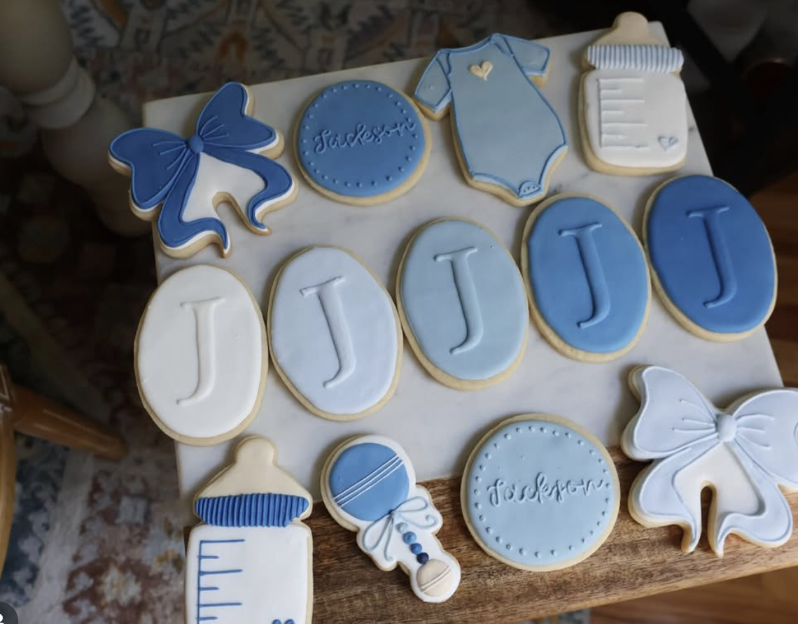 Decorated cookies for a baby shower or baptism, including a blue bow, baby onesie, baby bottle, rattle, and cookies with the name Jackson and the letter J, all in blue, white, and light blue icing.