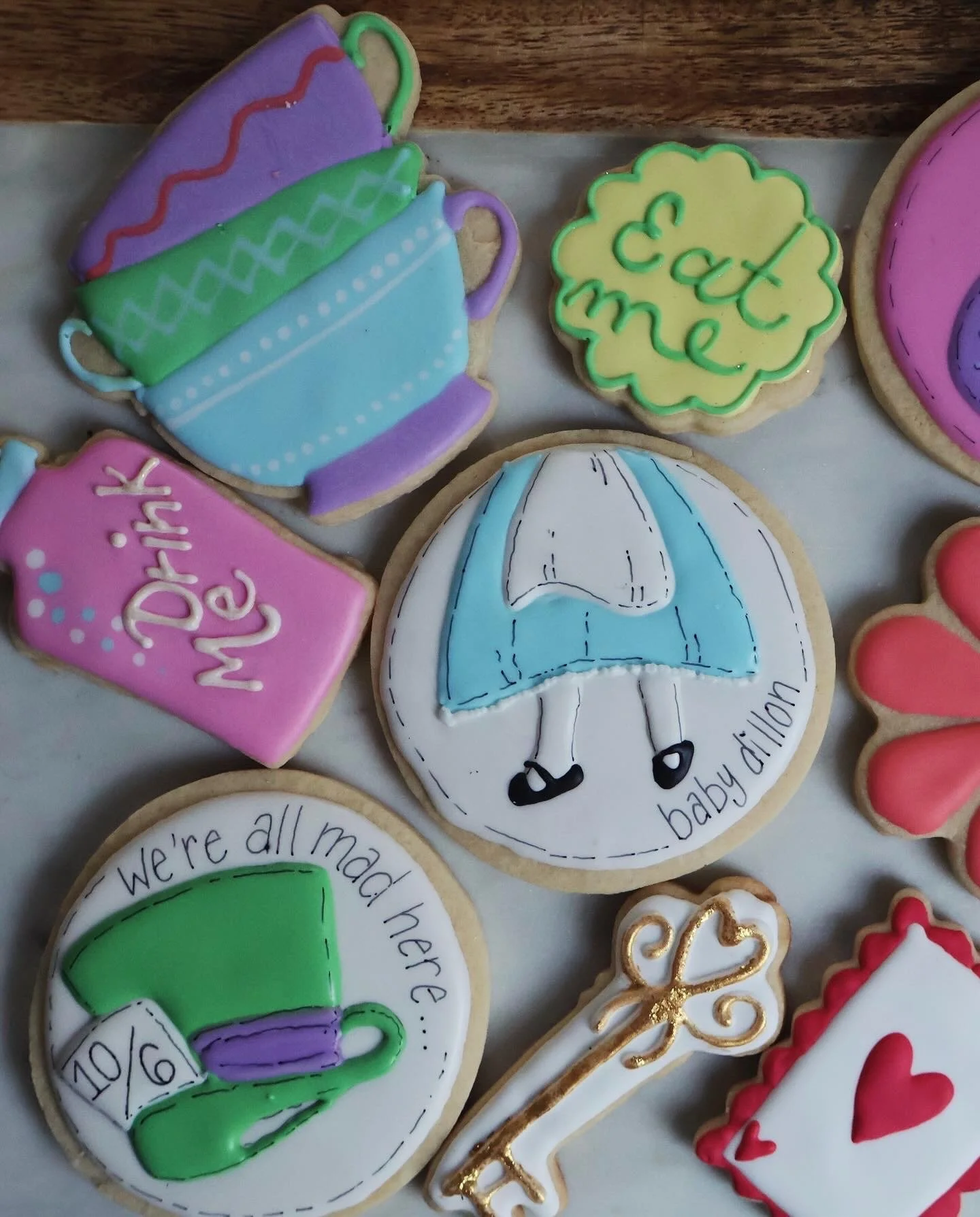 Decorated cookies with baby shower themes including a stack of colorful teacups, a yellow cookie with green icing and 'eat me' written, a pink cookie with 'Drink Me' written, a cookie with a blue umbrella and the words 'baby shower', a green and purp