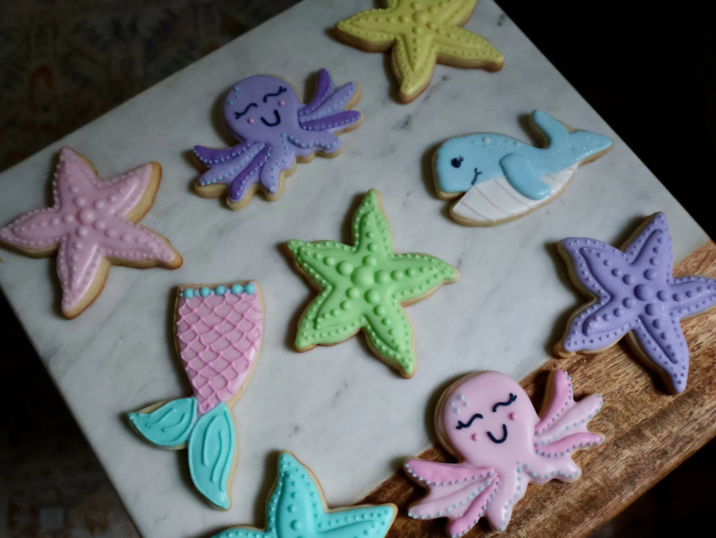 Decorated sugar cookies in ocean theme including pink, purple, and green starfish, a pink and purple octopus, a blue whale, a yellow starfish, a pink fish, and a purple starfish, arranged on a marble surface.