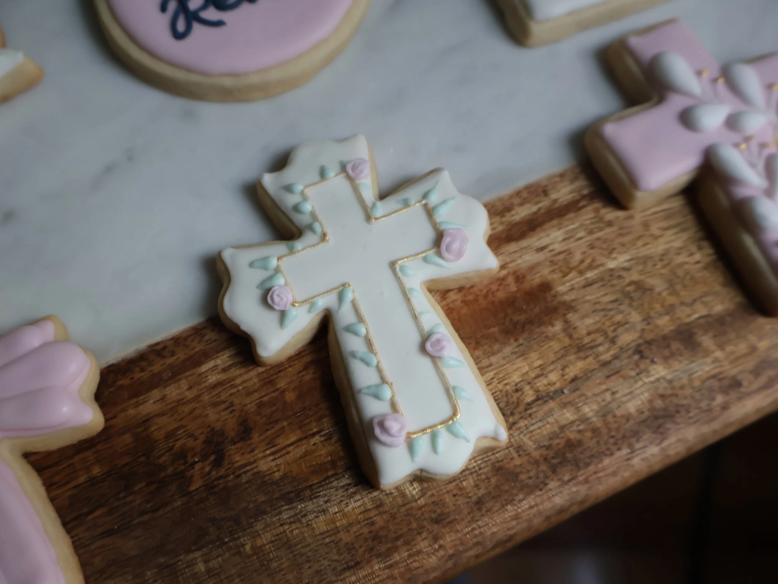 Decorated sugar cookie shaped like a Christian cross with white icing, pink and green floral decorations, and gold piping, placed on a wooden surface.
