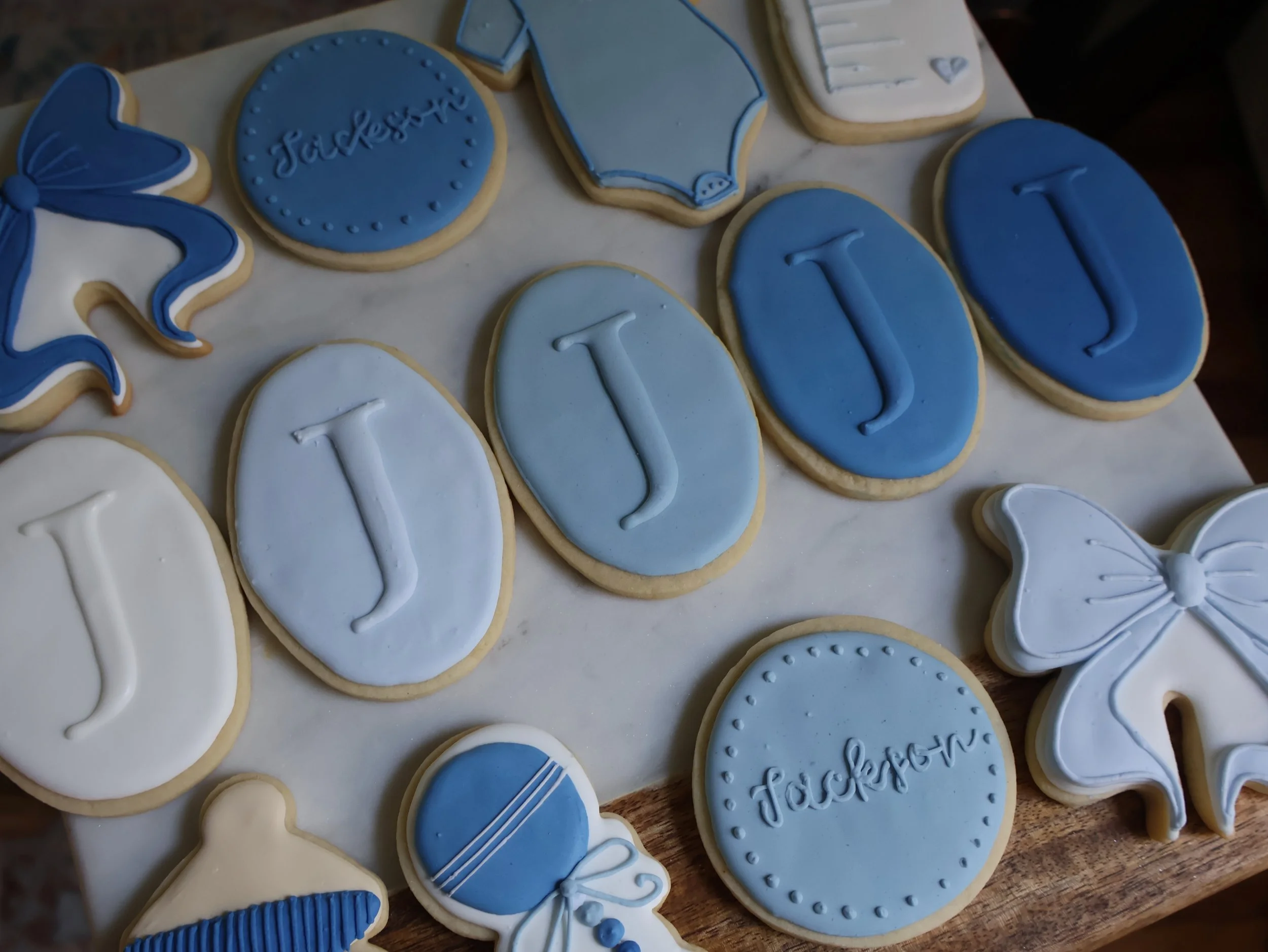Cookies decorated in blue and white with the letters 'J' on oval cookies, the name 'Jackson' on round cookies, and various other decorative cookies, including butterflies and a pacifier.