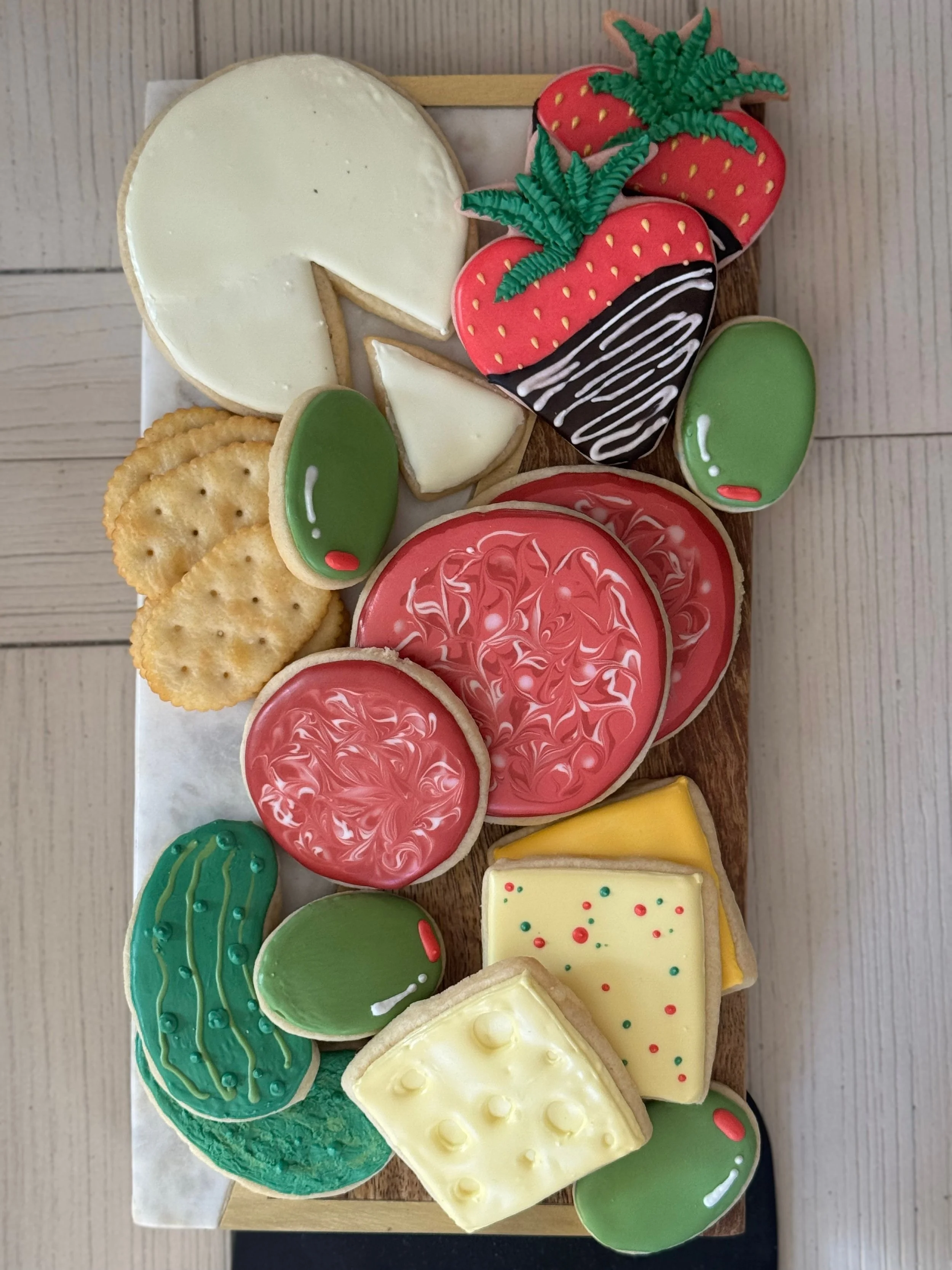 Assorted decorated cookies including strawberries, cheese, citrus slices, green vegetables, and round cookies arranged on a wooden tray.