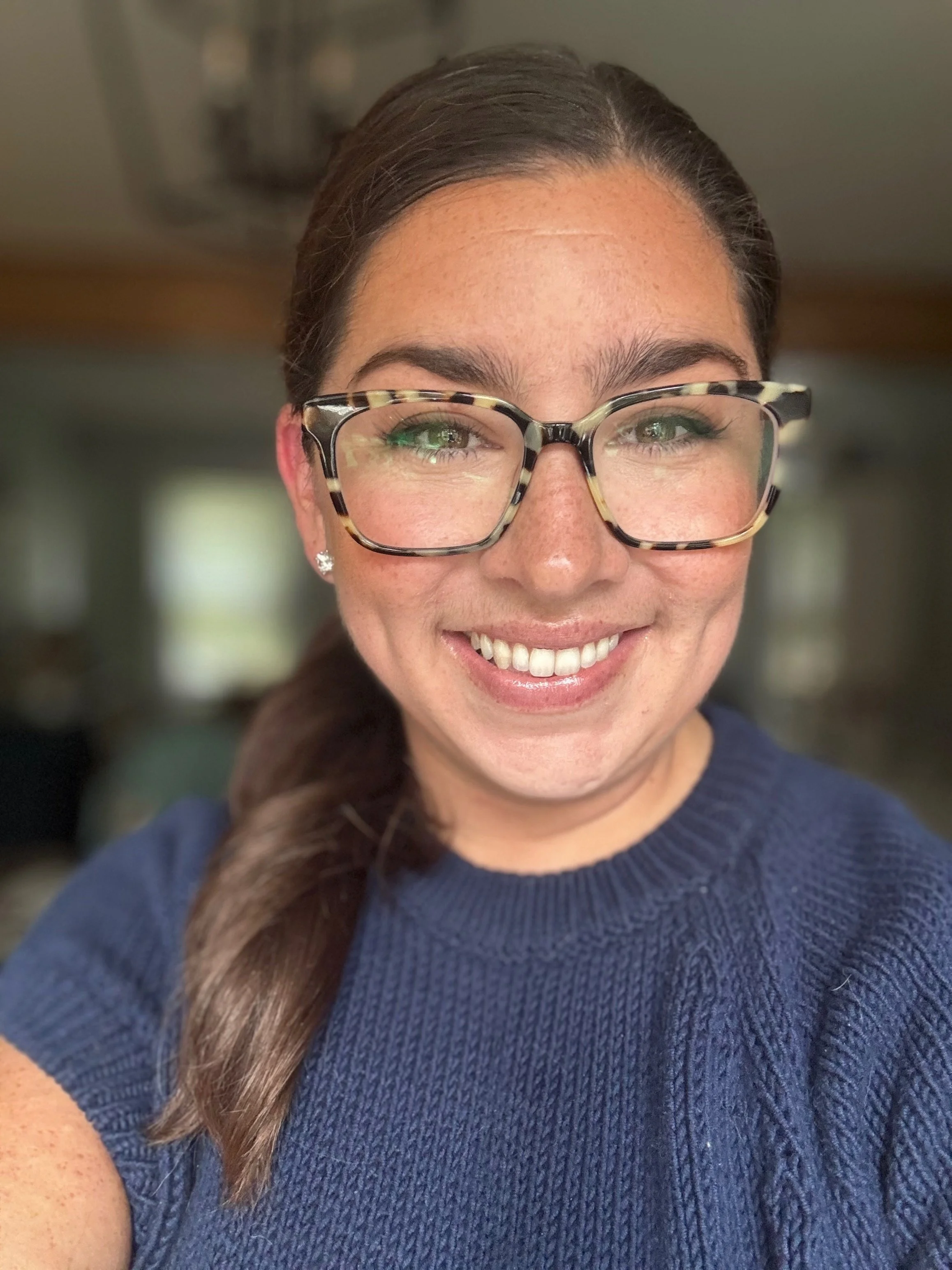 A woman with large glasses, brown hair tied in a side ponytail, and wearing a navy blue sweater, smiling in a well-lit room.