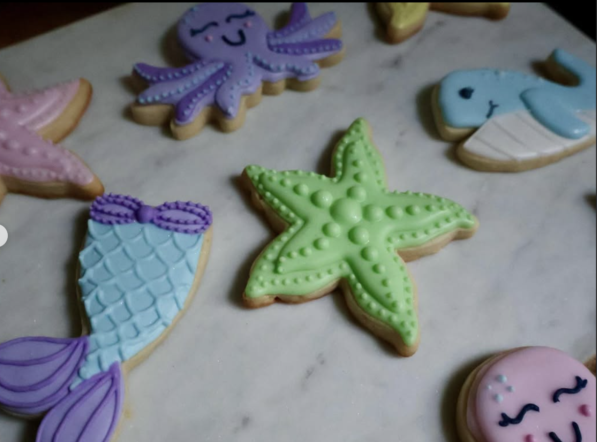 Assorted colorful ocean-themed decorated cookies, including sea stars, a whale, a mermaid tail, a purple octopus, and smiling pink and blue faces on a white surface.