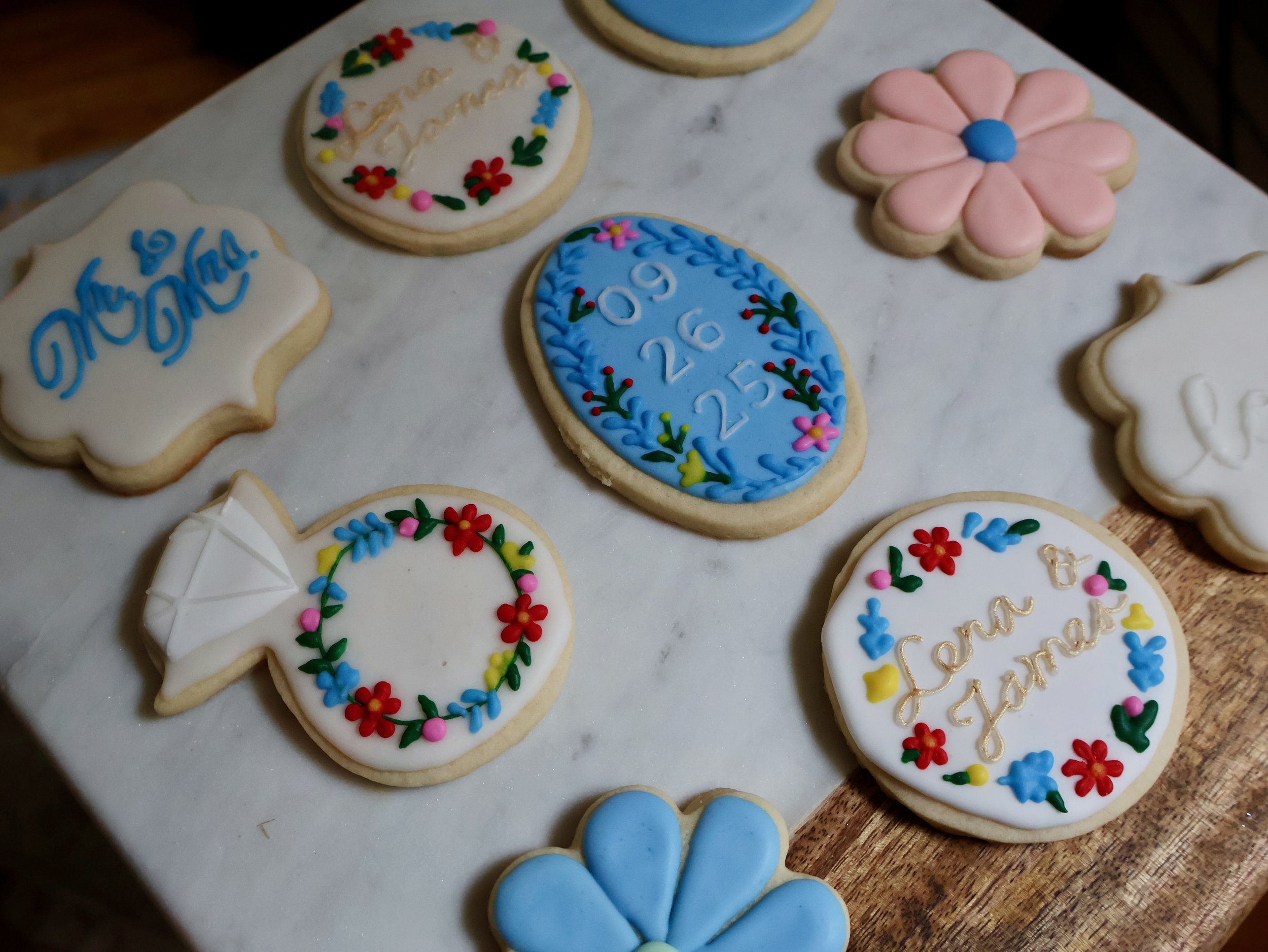 Decorated sugar cookies with floral and celebratory patterns, some with the name 'Cara Jeanne' and a date, '09 26 25', on icing, on a white marble surface.