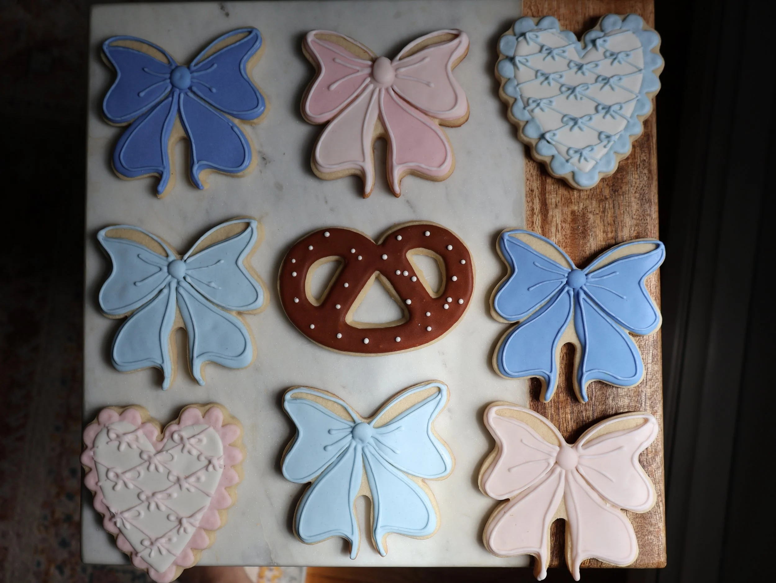 Decorated cookies on a wooden and marble surface, including six butterfly-shaped cookies in blue, pink, and white with pastel icing, two heart-shaped cookies with pink and white icing, and a pretzel-shaped cookie with brown icing and white dots.