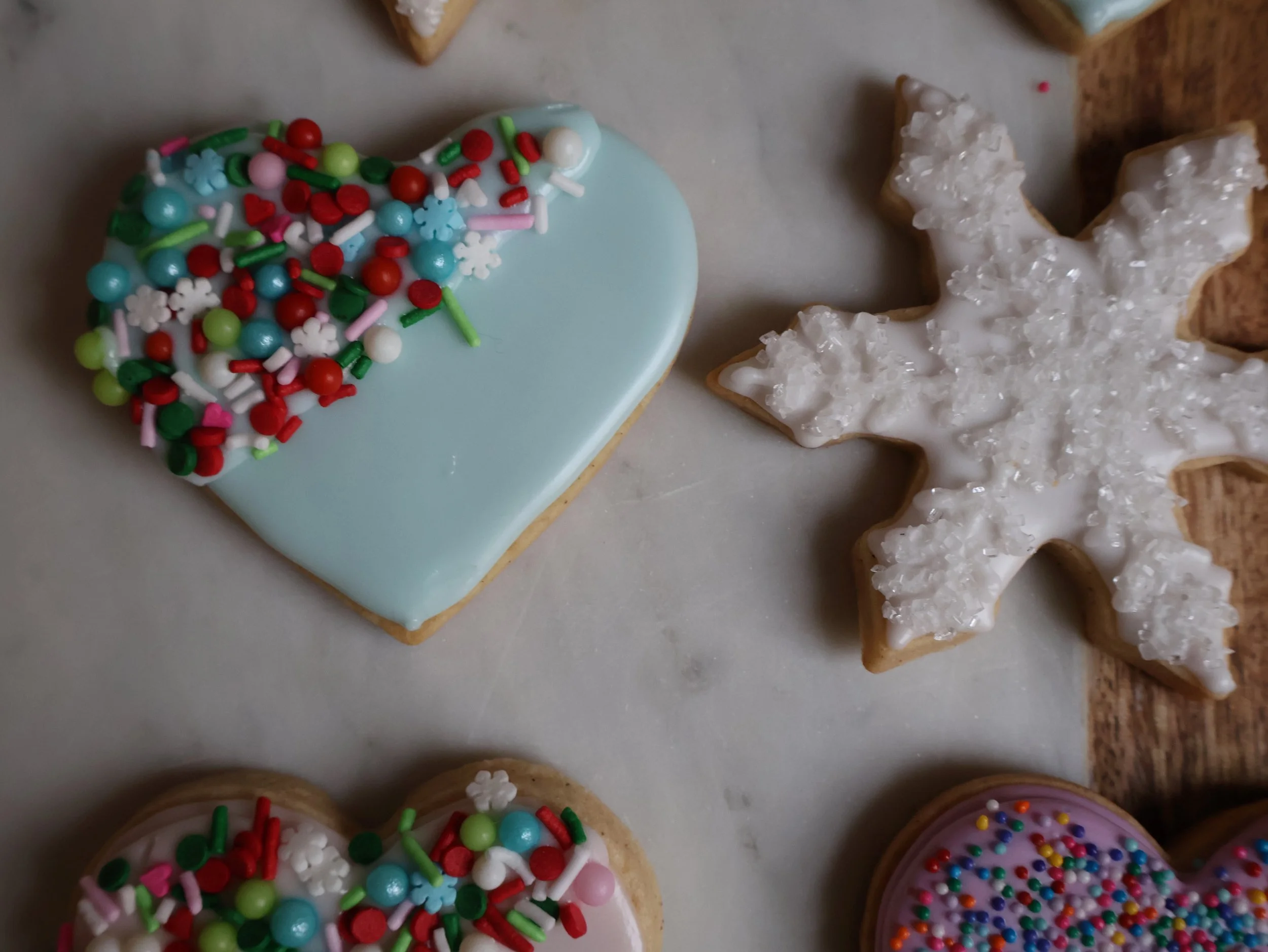 Decorated Christmas cookies on a marble surface, including a heart-shaped cookie with light blue icing and sprinkles, and a snowflake-shaped cookie with white icing and sugar crystals.