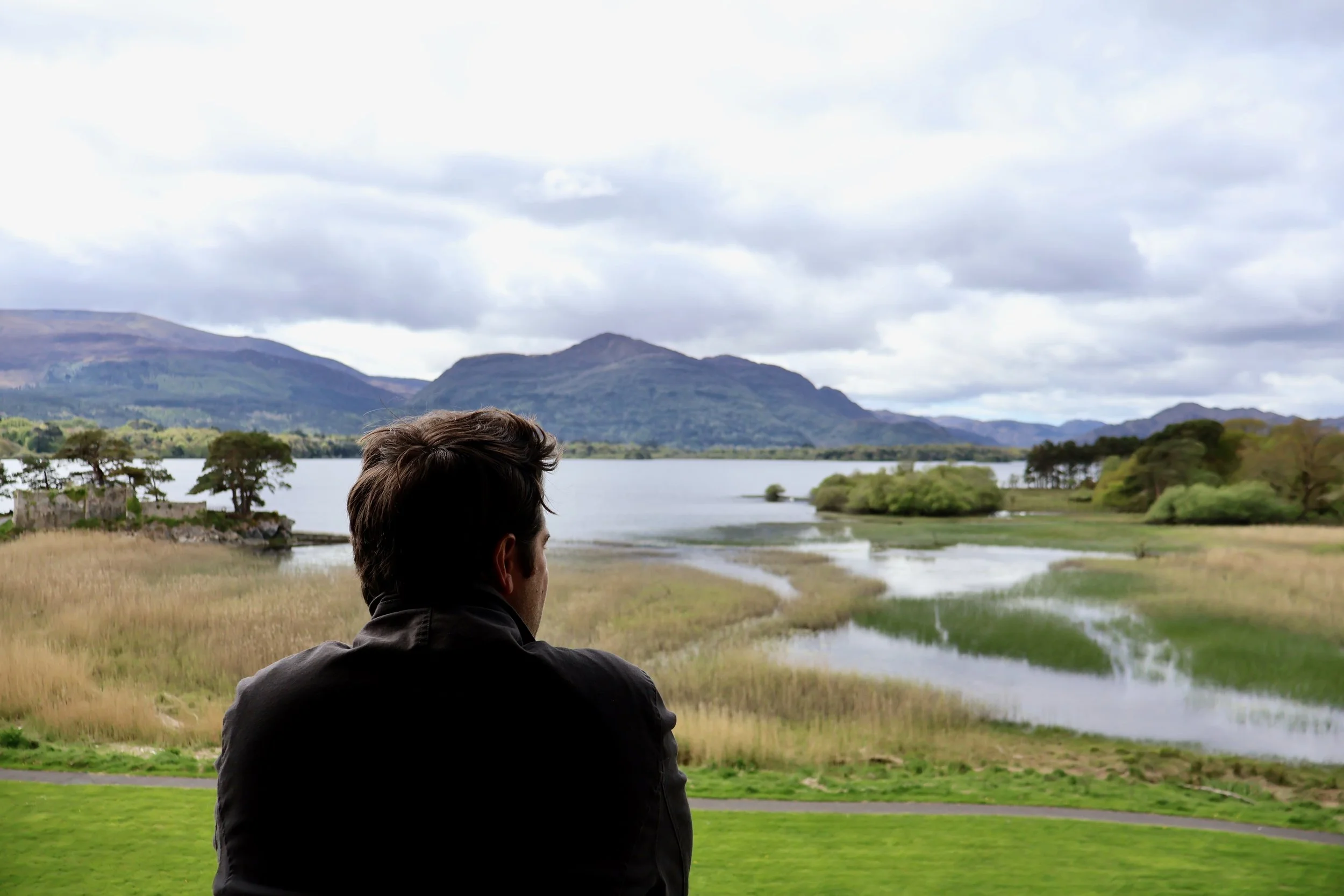 A man with dark hair sits on a grassy area, looking out at a lake surrounded by mountains and trees under a cloudy sky.