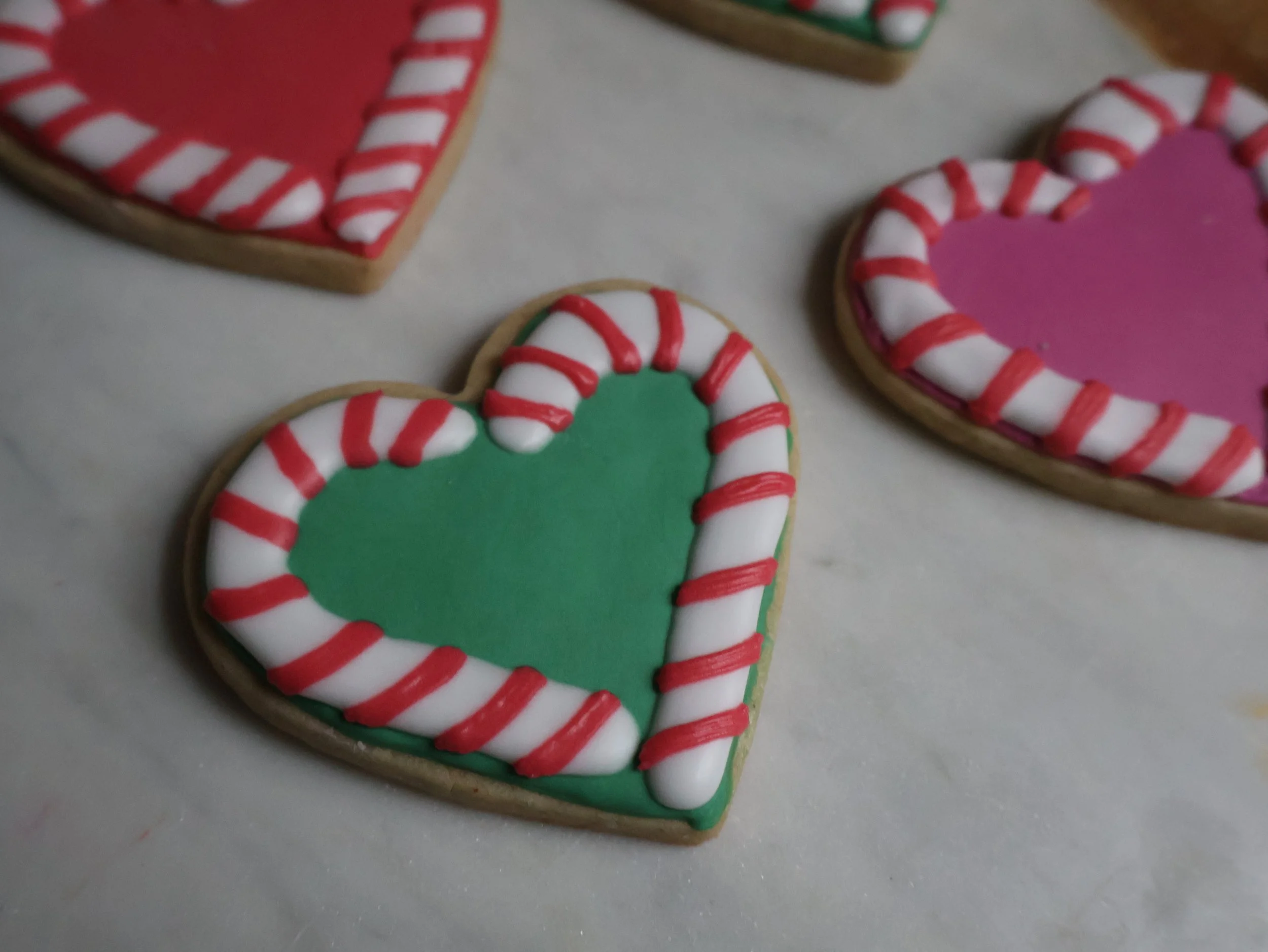 Decorated heart-shaped cookies with green and pink icing and red and white striped candy cane borders on a white surface.