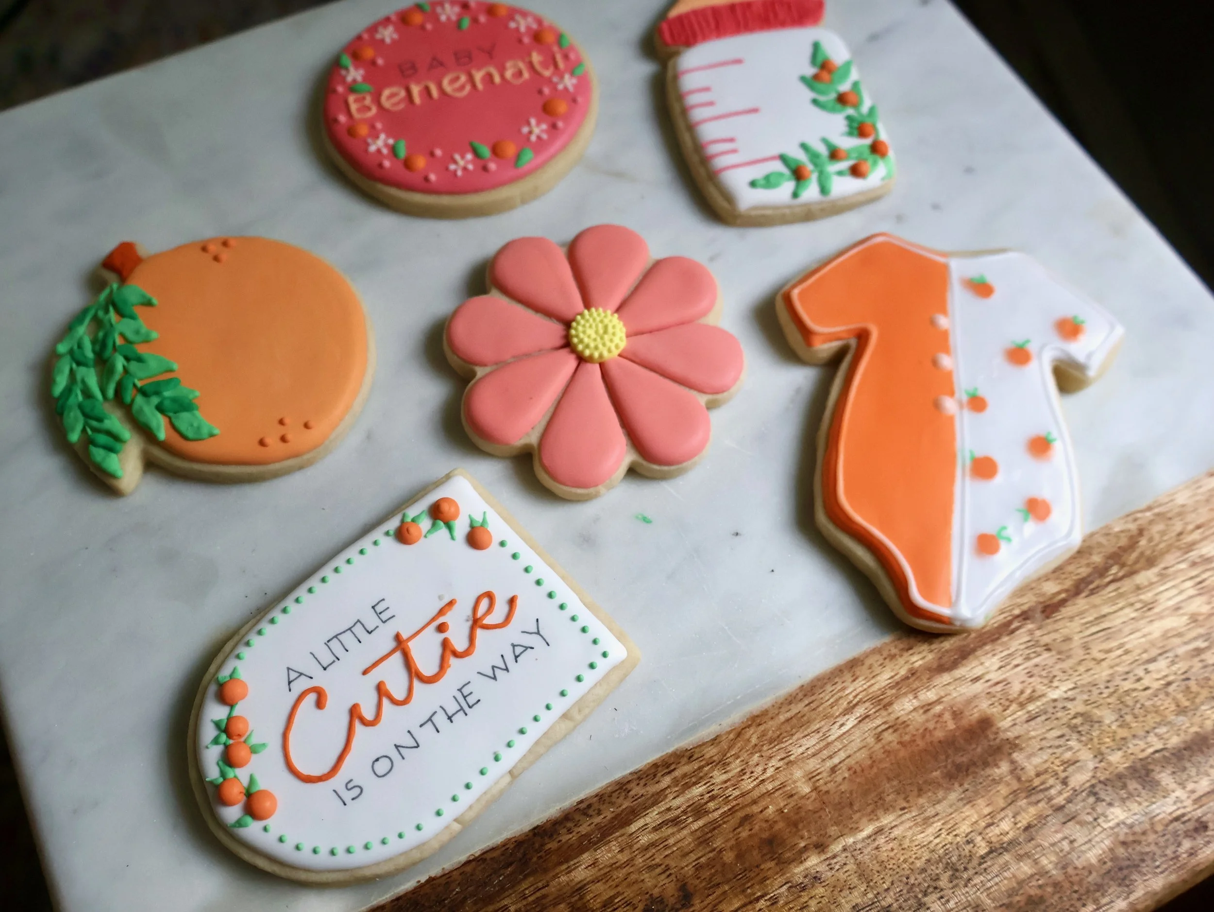 Decorated cookies in festive shapes and designs, including a pumpkin, pink flower, orange and white pumpkin, measuring tape, a pink circle with the words 'Baby Benedict', and a white rectangle with the phrase 'A little Cuter is on the way'.