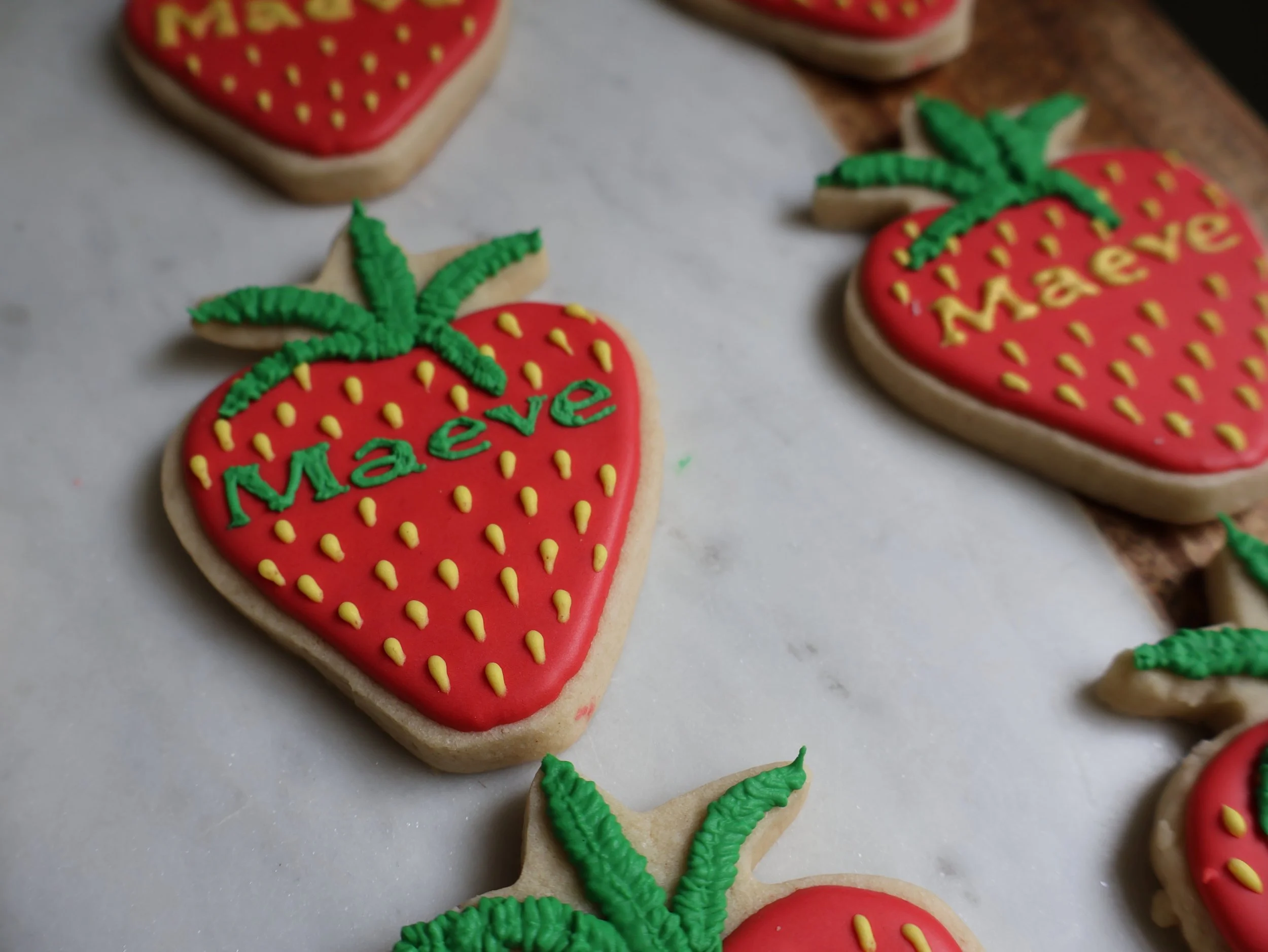 Decorated sugar cookies shaped like strawberries with red icing, green leaves, and the word 'Maaeve' written in yellow and green icing.