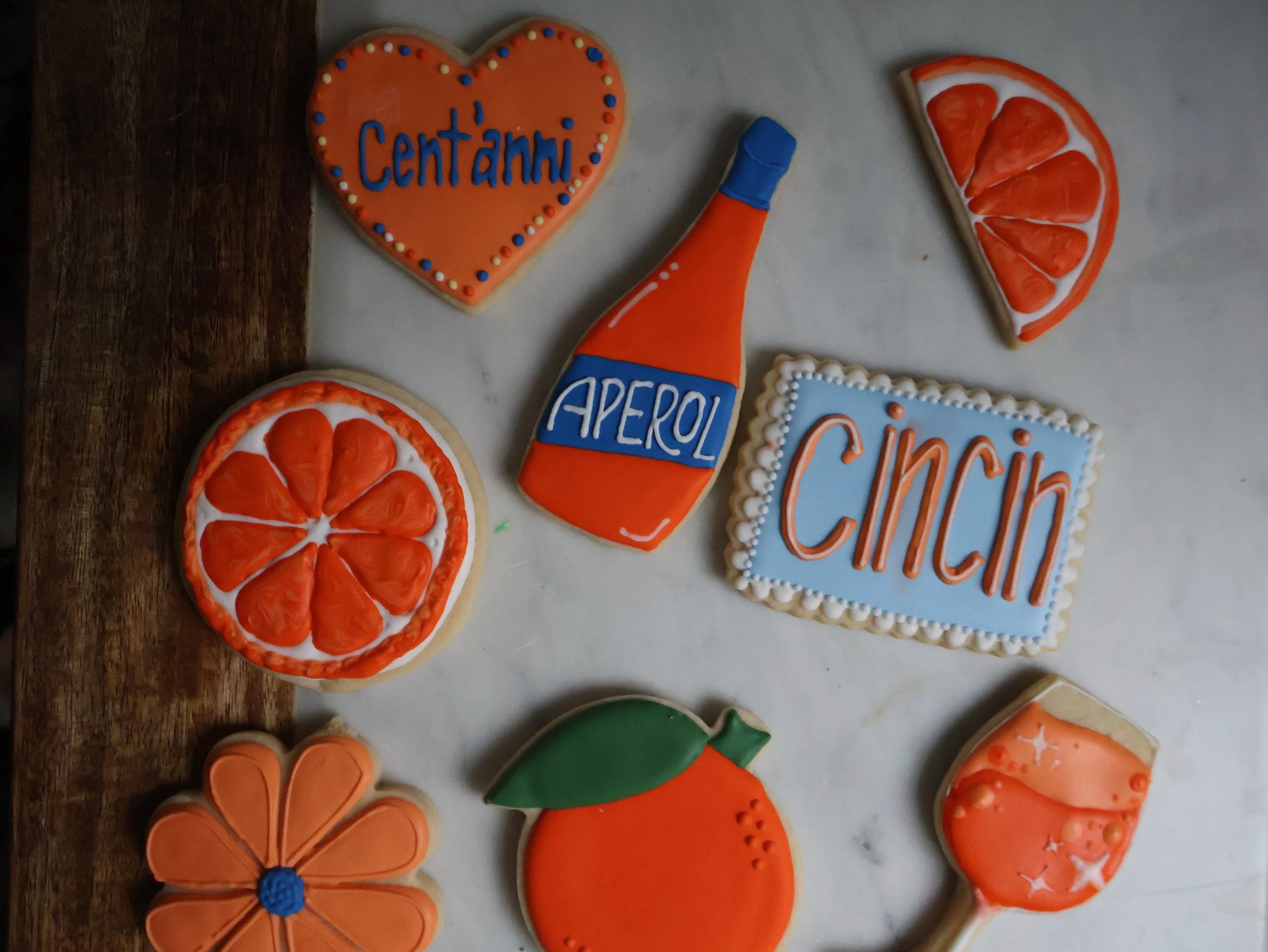 Decorative cookies shaped and decorated like orange slices, a heart with 'Centanni' written, a bottled beverage labeled 'Aperol', a square with 'Cinci' written, an orange fruit, a flower, and a cocktail glass with a drink.