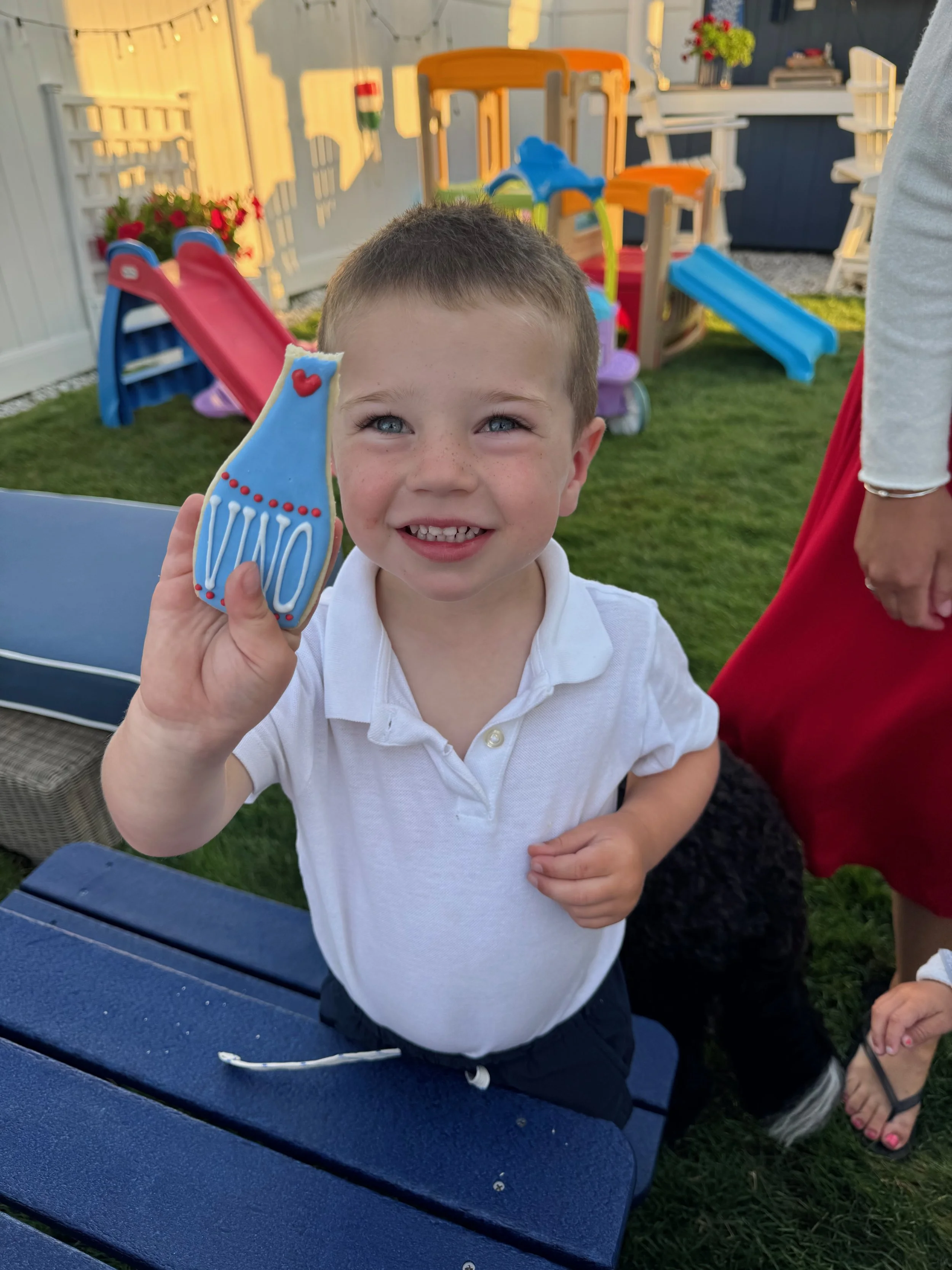 Young boy smiling and holding a decorated cookie shaped like a sock or stocking at an outdoor birthday party with playground equipment and colorful decorations in the background.