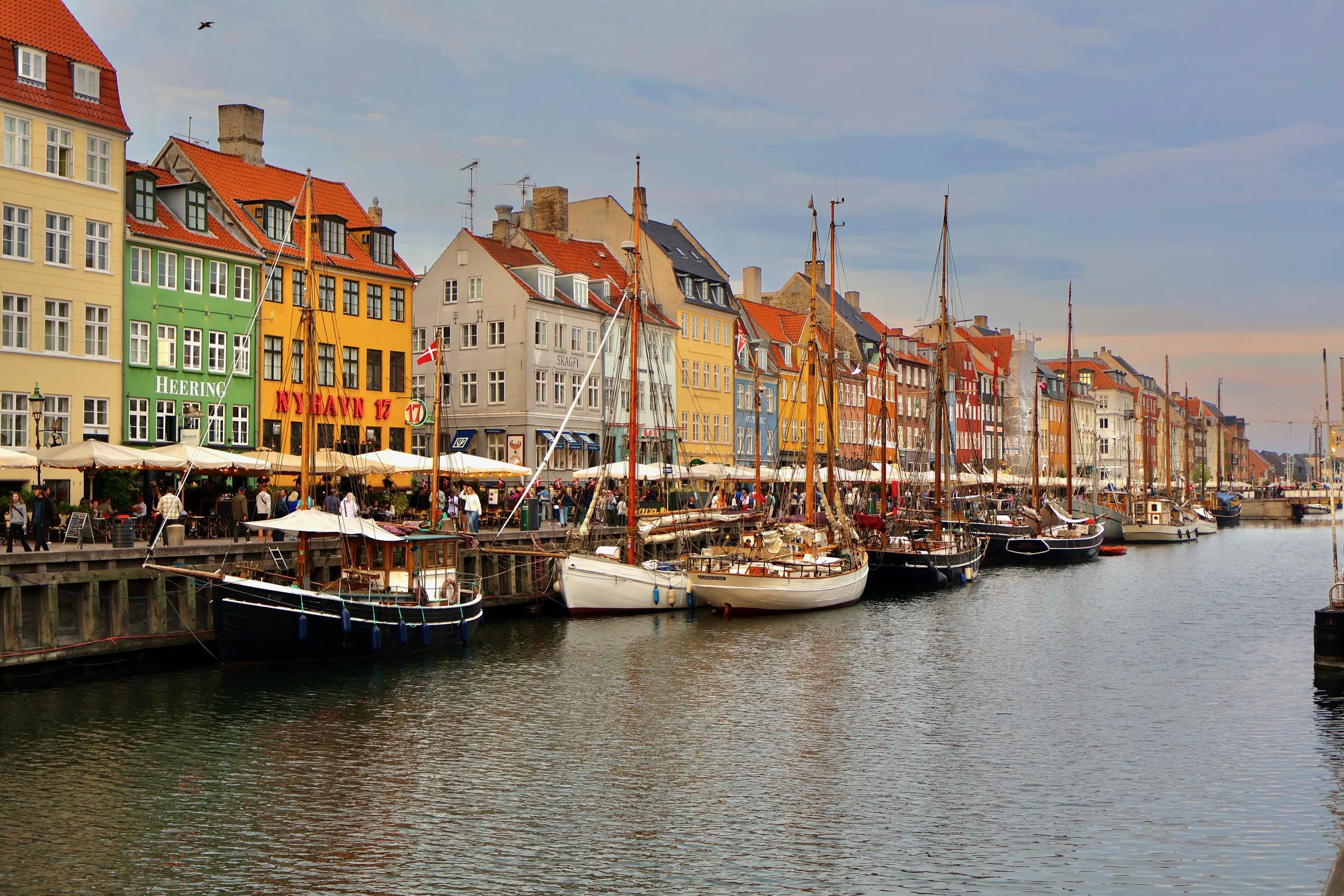 Colorful buildings and sailboats docked along a harbor with outdoor cafes and people walking.