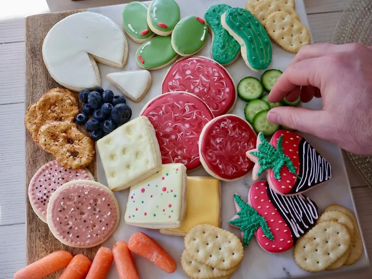 Decorative cookies shaped like strawberries, strawberries with leaves, and cakes with striped and polka dot icing, arranged on a white platter with cucumber slices, blueberries, carrots, and assorted crackers.