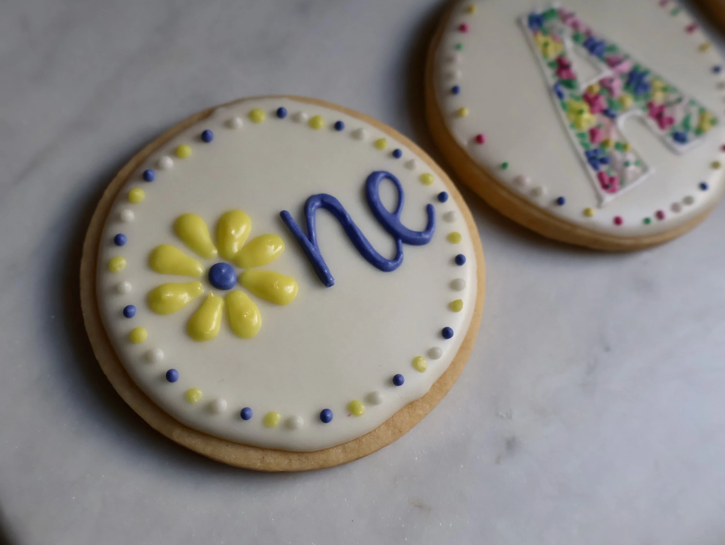 Decorated cookies with white icing, yellow and purple sprinkles, one has yellow flower and the word 'Love' written in blue, the other has a colorful butterfly design.