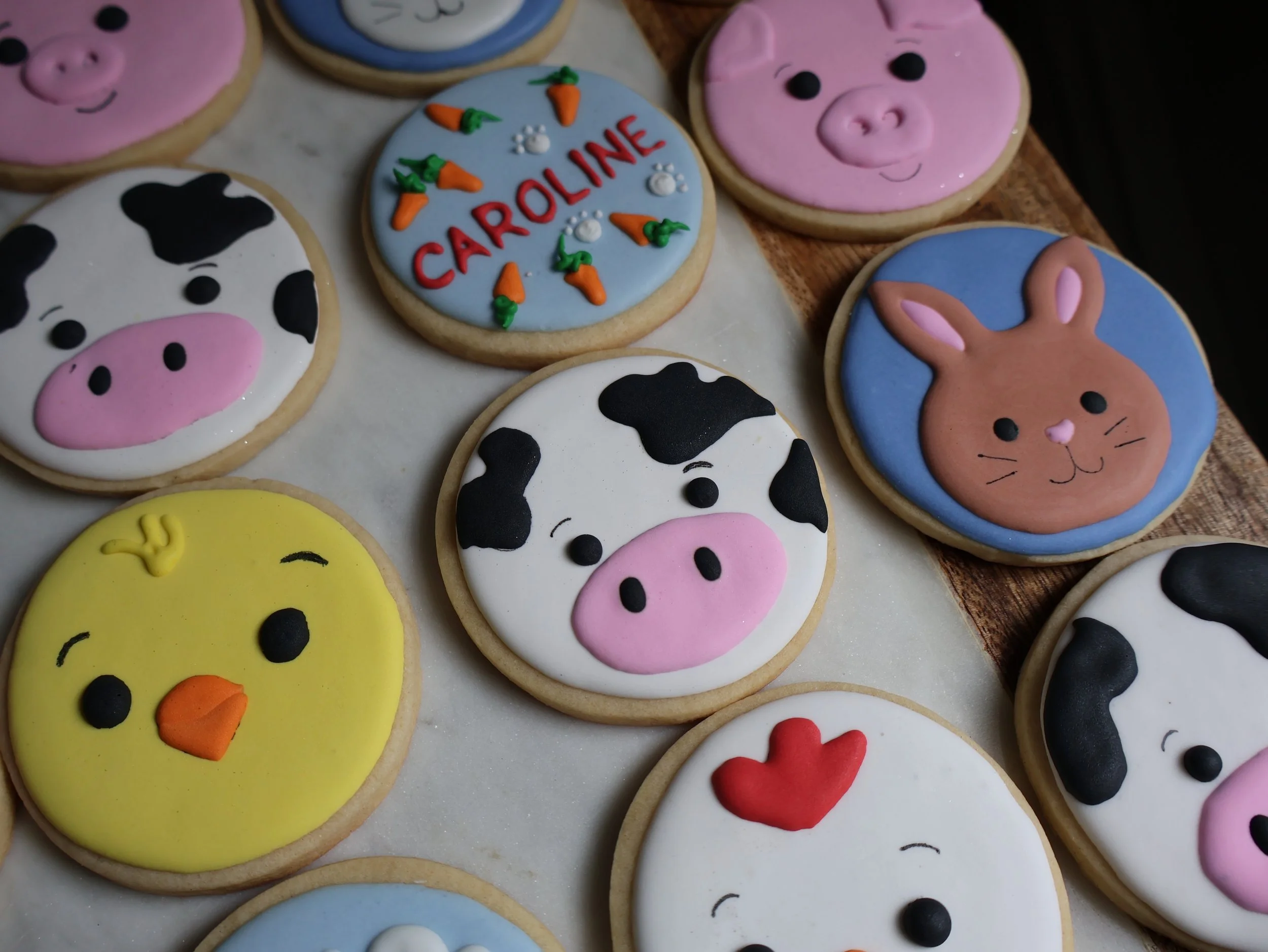 Decorated cookies with farm animal faces, carrots, and the name 'Caroline' on a blue cookie, placed on a wooden surface.