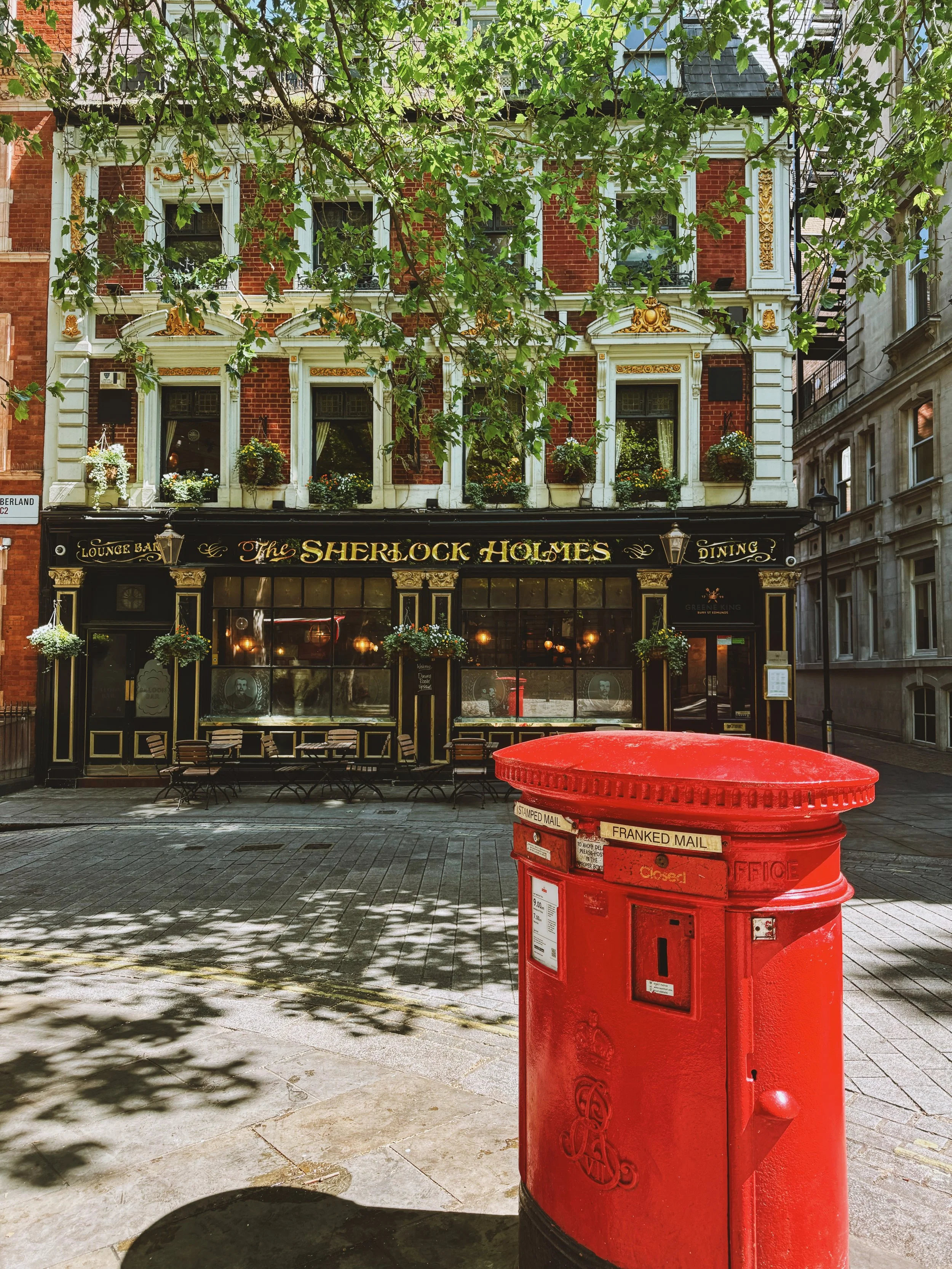 Red British mailbox on a cobblestone street in front of a building with a pub called 'The Sherlock Holmes'.