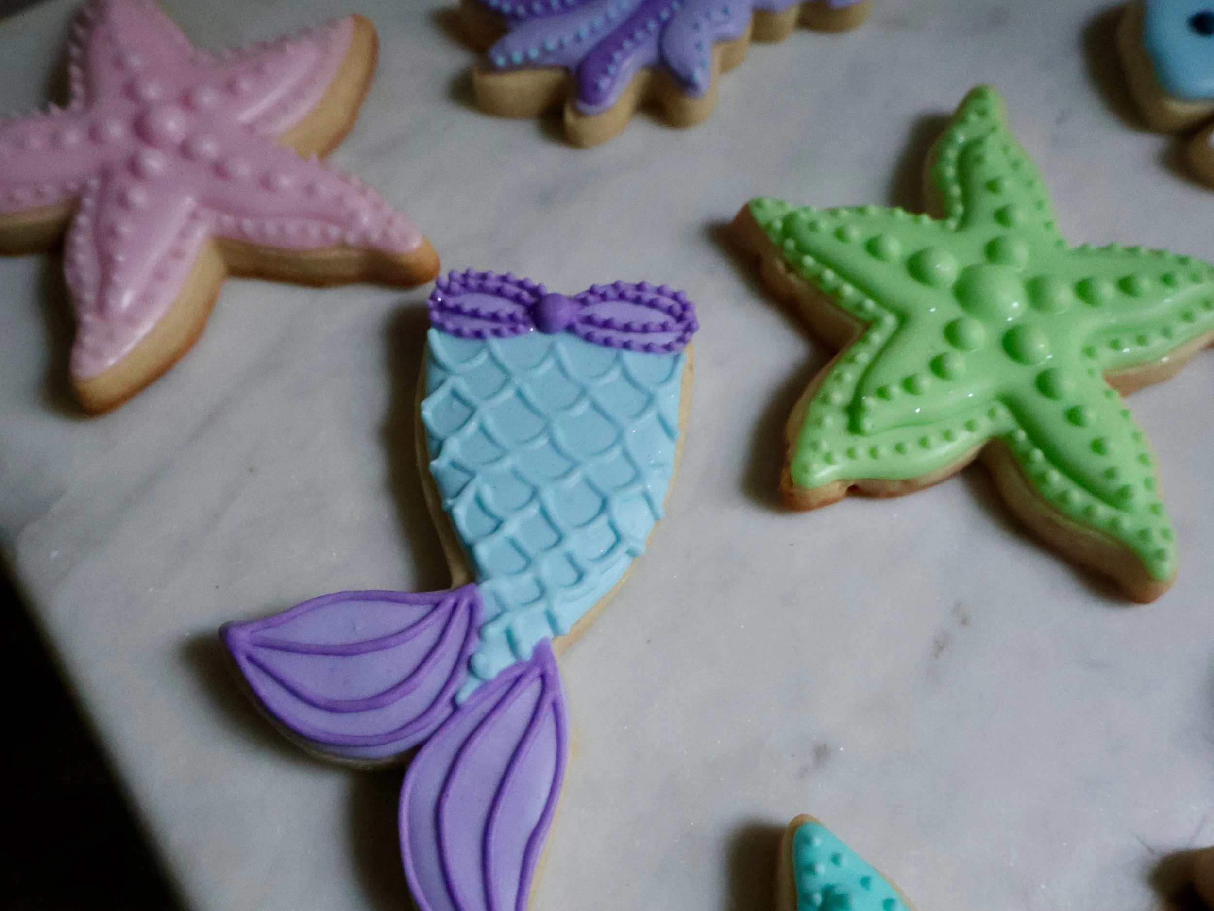 Colorful starfish and mermaid tail cookies decorated with pastel icing on a white surface.